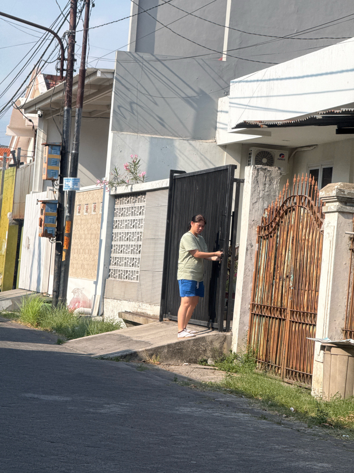 In Surabaya, Indonesia, a person stands near a black metal gate, reaching for its handle. They are wearing a green t-shirt and blue shorts. Adjacent to the black gate is a rusted, ornate metal gate. The area appears to be a residential street with houses visible in the background, including a white building with an air conditioning unit. Overhead, a complex network of electrical wires crisscrosses the scene. The lighting suggests it is daytime, with shadows cast on the asphalt road. A grey trash bin is situated next to the rusted gate.