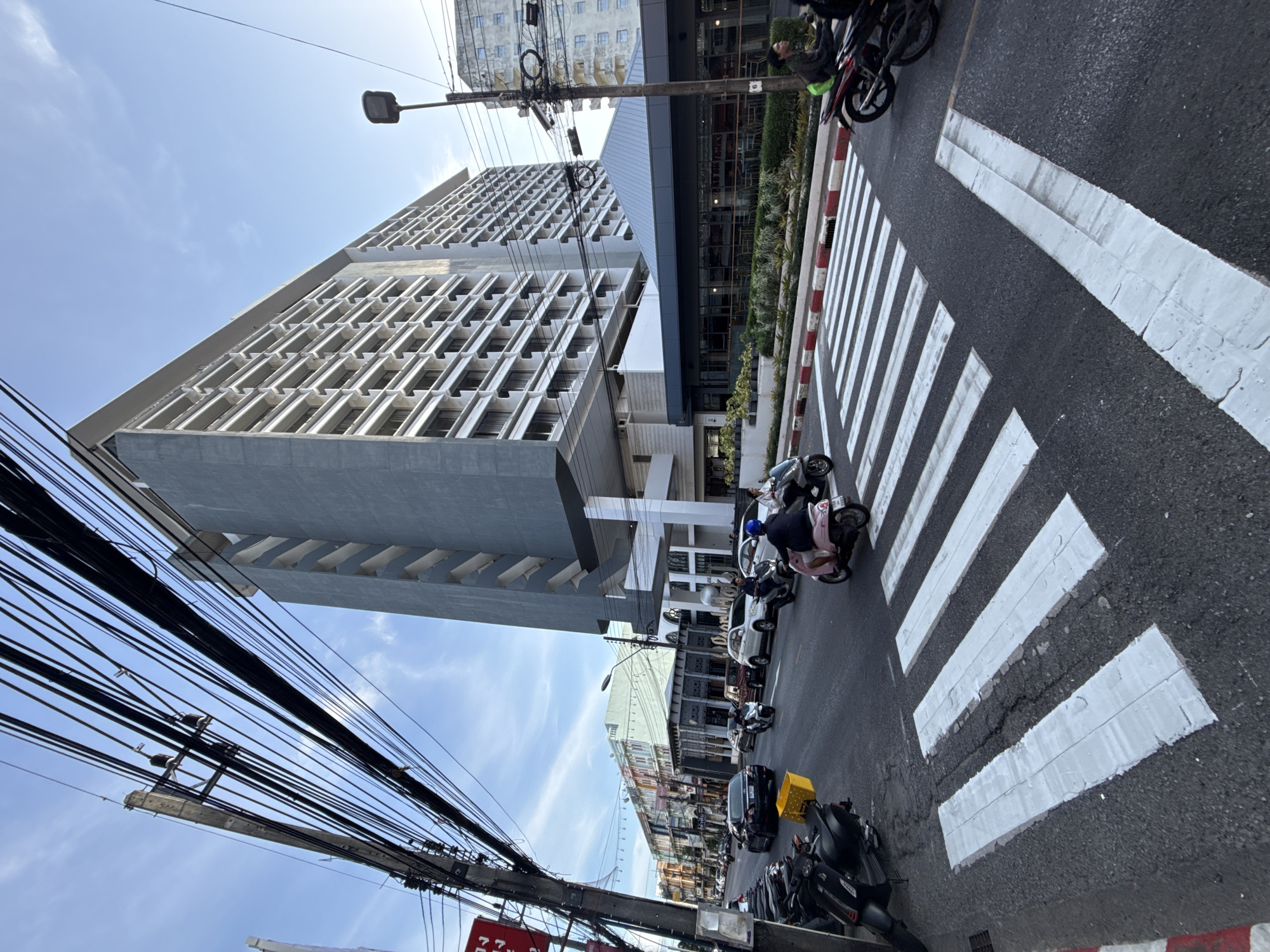 A wide-angle, eye-level shot captures a busy street scene in Phuket City Municipality, Thailand, on a clear day. In the foreground, a zebra crossing dominates the asphalt road, marked by thick, white stripes with a red and white border. Multiple motorcycles are parked along the left side of the street, with a yellow bin attached to the back of one.
A procession of motorcycles is actively navigating the road and crossing the pedestrian path. One rider, wearing a helmet, is on a pink scooter, while another, on a black scooter, is behind. Several other motorcycles and a white SUV are also visible in motion.
The street is lined with buildings. To the right, a tall, modern, grey building with numerous balconies and windows rises majestically, its facade punctuated by stairwell structures on its left side. Below this structure, a darker, glass-fronted building with outdoor seating and greenery is situated. Further down the street, a row of colorful, multi-story buildings is visible, suggesting a commercial or residential area. Overhead, a dense network of electrical wires crisscrosses the sky, supported by a wooden pole on the left. A streetlamp is also present on the right side of the frame. The sky above is blue with scattered clouds.
