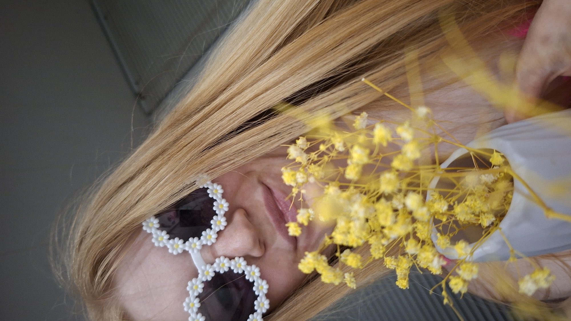 The scene is set in the city of Chaikovsky, Russia. A female subject with long, straight, golden-blonde hair is depicted from a low-angle, close-up perspective. She wears white-framed sunglasses adorned with small, five-petaled white flowers that have yellow centers, resembling daisies. The dark lenses of the sunglasses obscure her eyes. Her lips are closed in a slight, soft smile. She holds a small, light grey, irregularly shaped vase containing a dense cluster of small, bright yellow dried flowers, consistent with dyed baby's breath. The flowers are positioned prominently in the foreground, slightly out of focus, and partially obscure the lower portion of her face, including her chin and mouth. A glimpse of a bright pink garment is visible on the lower right side of the frame, underneath her hair. The background features muted grey tones, with a uniform surface above and a textured, possibly ribbed or paneled grey surface to the lower left.