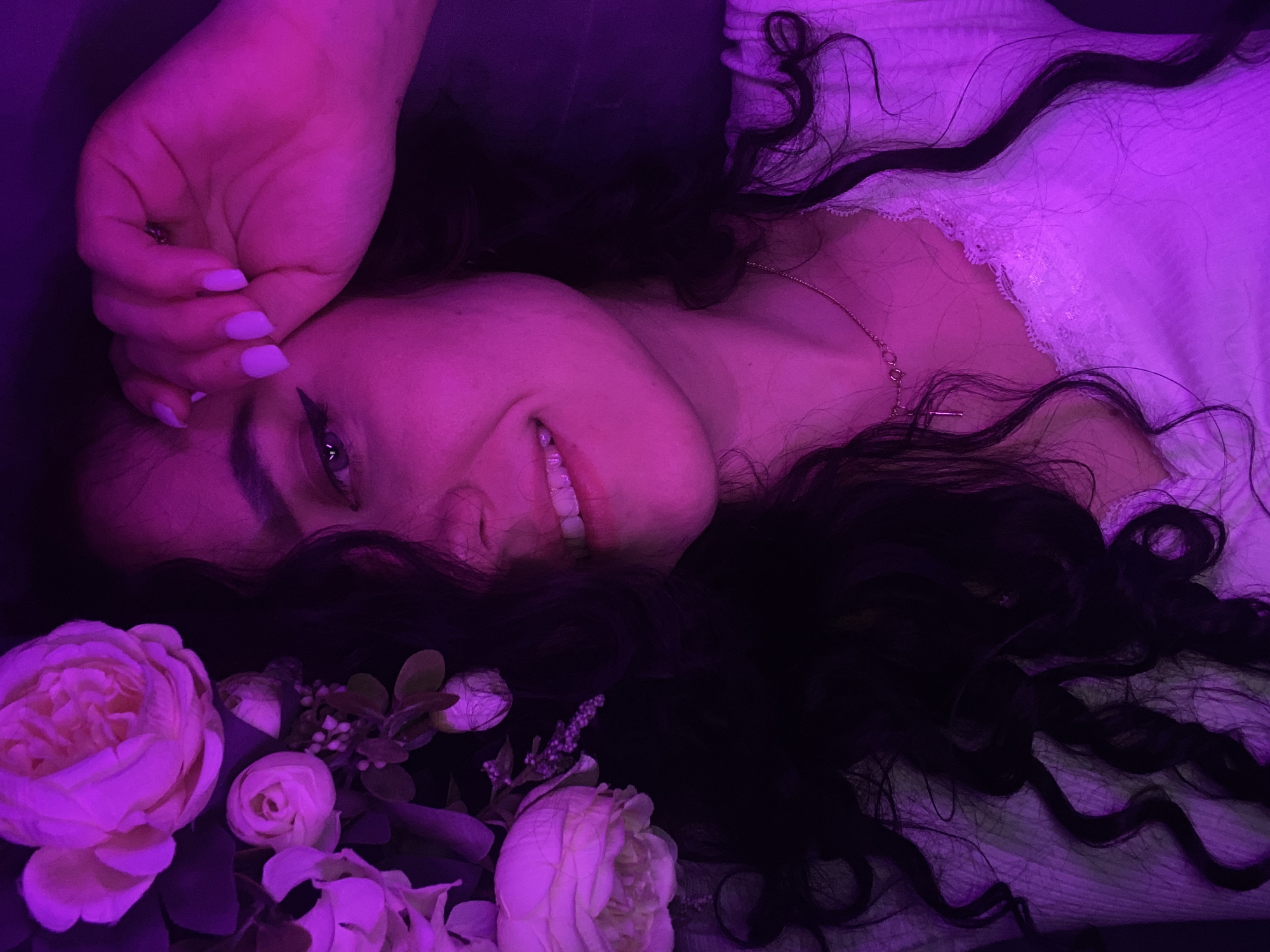 A woman lies on her back amidst light-colored flowers. She has dark, curly hair and is wearing a light-colored top with lace detail. Her left hand is raised near her face. The image is lit with a purple light, creating a distinct hue over the entire scene.
There is no information about the location in the city of Chaikovsky, Russia in the image.