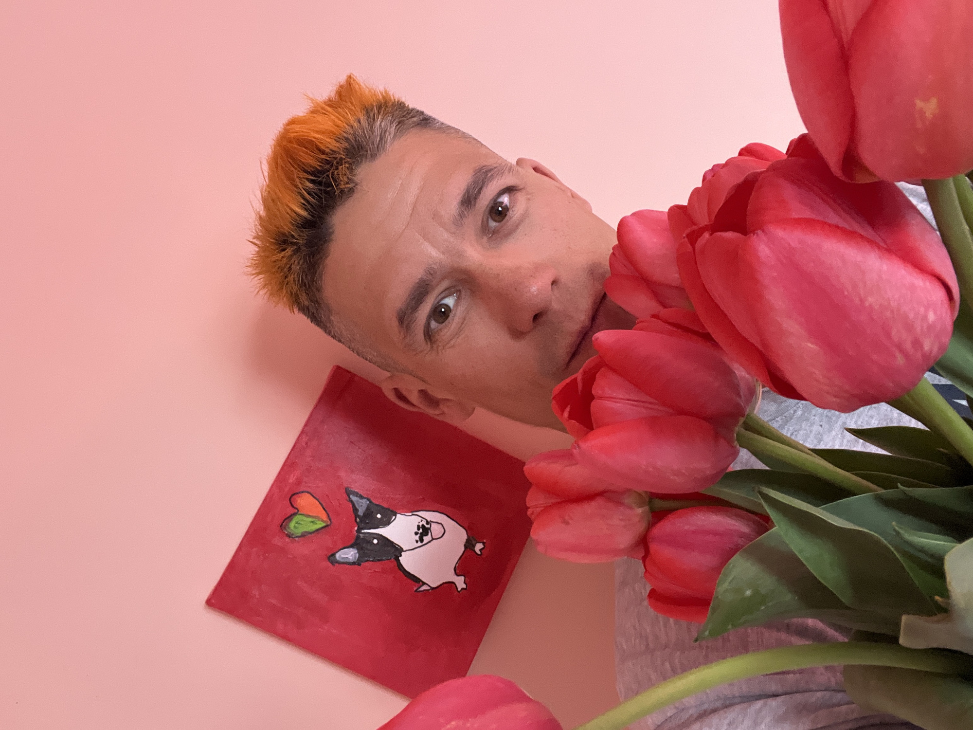 A close-up, slightly angled photograph features a man with short, dyed orange hair, light skin, and brown eyes, posing with a bouquet of red tulips. He is looking into the camera. Next to the man is a red painting with a drawing of a black and white dog with heart-shaped ears. The background is a pale pink color. The scene is likely indoors. No specific details confirm its location in the city of Chaikovsky, Russia.