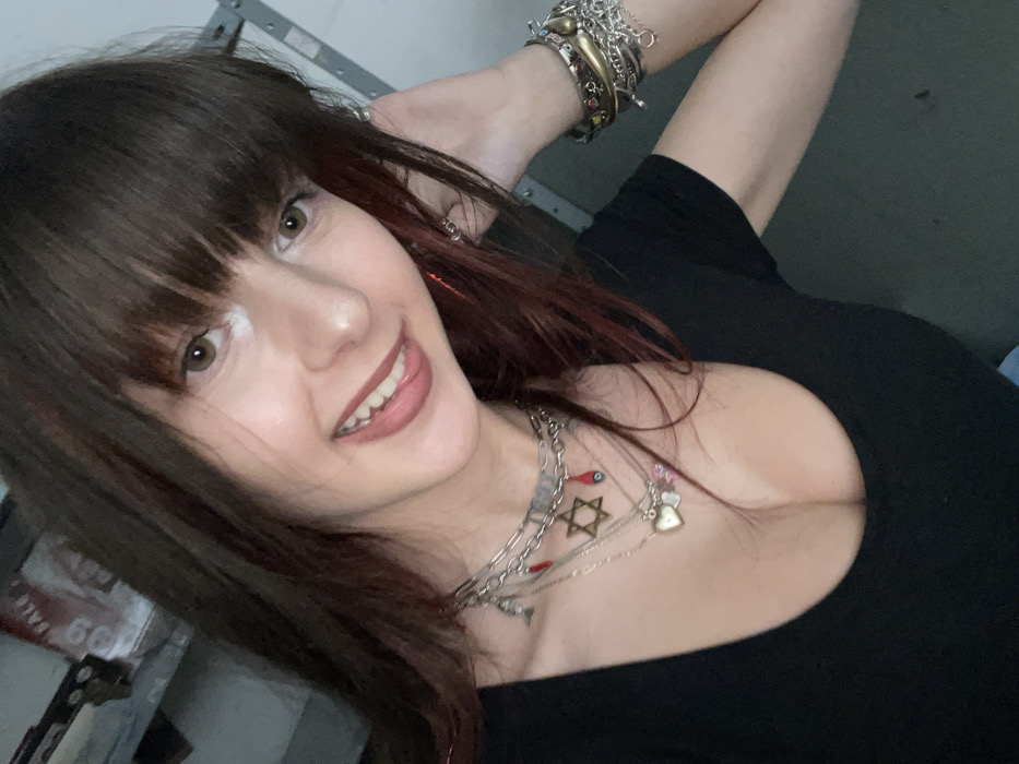 The image displays a close-up portrait of a young adult female, framed from the chest up, positioned indoors. She has long, dark brown hair with subtle red streaks and straight bangs. Her complexion is light, and she exhibits a smiling expression with her teeth visible and the tip of her tongue slightly extended. A distinct white mark is present on the bridge of her nose. She wears a black, short-sleeved top with a low scoop neckline. Her neck is adorned with multiple layered silver-toned necklaces; one features small red chili pepper charms and an eye charm, another displays a Star of David pendant and a red evil eye charm, and a third includes a heart-shaped locket with smaller charms. Her left wrist (viewer's right) is covered with a dense stack of various silver-toned bracelets. Her left hand is raised, with fingers touching her hair near her temple. The background is out of focus but appears to be an interior space, possibly a utility area or garage, featuring metallic shelving and a partially visible white sign with red lettering, including the number '99', on the upper left. Metallic vertical structures are also discernible on the right side of the background. The scene is situated in Raanana, Israel.