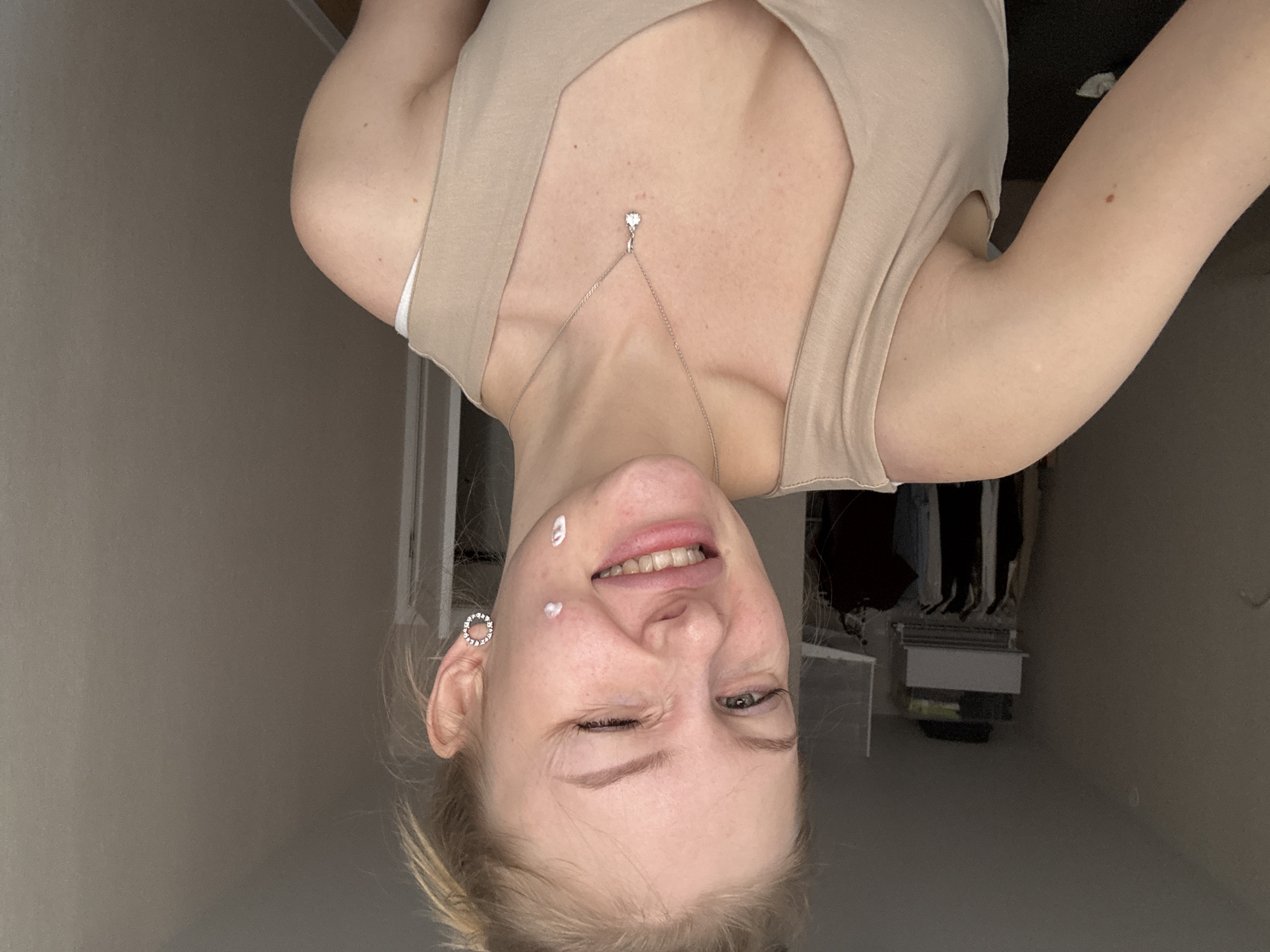 A woman is pictured in an indoor setting, wearing a light beige top and a delicate necklace with a pendant. She is facing the camera with her head tilted upside down and a playful expression, winking her left eye. Two small dots of what appears to be cream are visible on her face: one on her cheek and another near her lip. Her right ear is adorned with a round earring. In the background, there is a glimpse of a closet containing hanging clothes. The setting is a room with neutral-toned walls. There is no information to support that this is in Perm, Russia.