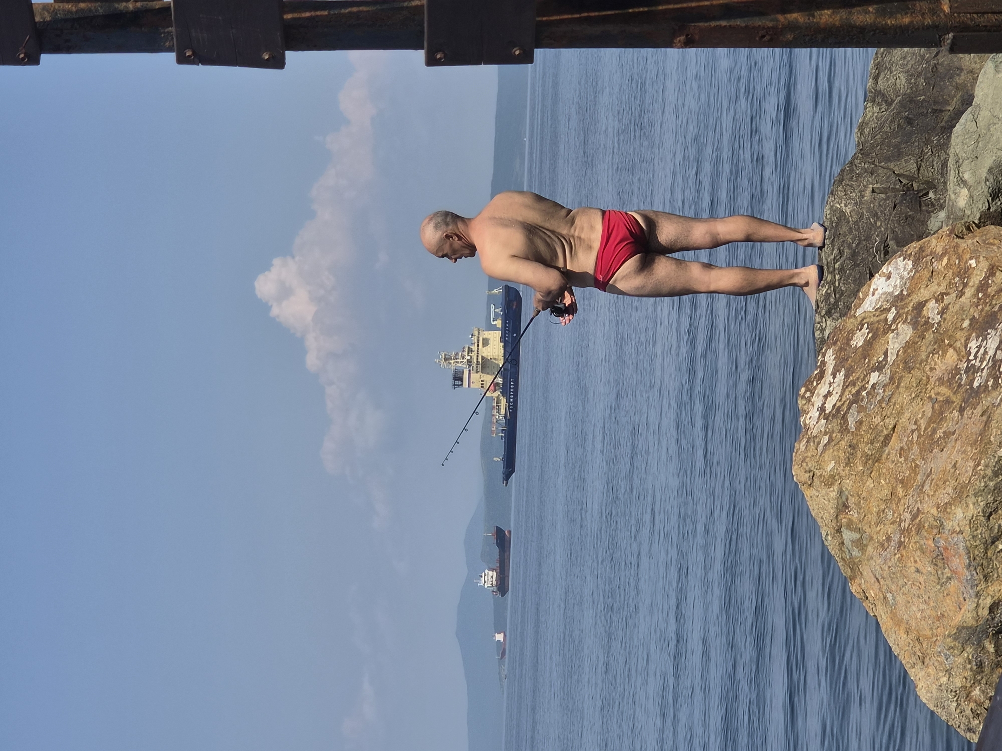 A bald, middle-aged man wearing red swim briefs stands on a rocky outcrop near the water and fishes. He is facing away from the viewer, looking out at the sea. In the background, several ships are visible on the water, and mountains can be seen in the distance. The sky is blue with a few white clouds. The scene appears to be a coastal area in Vladivostok, Russia.