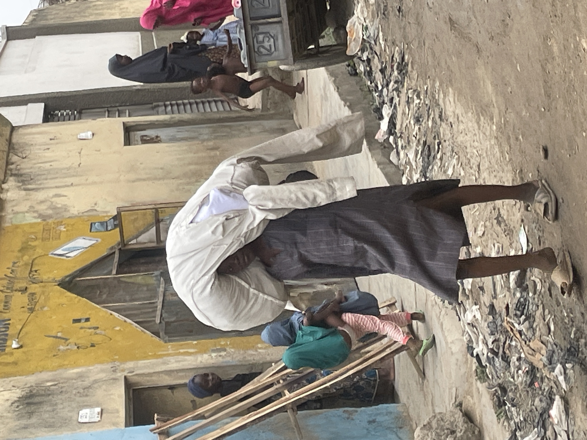 An outdoor daytime scene depicts a man carrying a large, bulky white sack on his back, walking along a dirt road in an urban environment. He wears a long, dark-colored robe. The road edges are littered with refuse. In the background, a yellow building is visible, with weathered walls and a partially constructed or dilapidated roof. Other people are present, including a child in the midground and other individuals further in the distance. Laundry hangs on a clothesline overhead. The location is in the city of Unknown, Unknown.