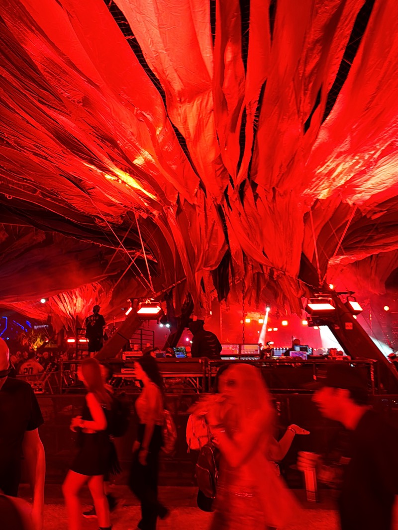 The image depicts an event, likely a music festival, occurring in Indio, United States, given the overall aesthetic and ambiance associated with events like Coachella.
The environment is bathed in red light, casting a stark and somewhat surreal atmosphere. Overhead, draped fabric resembling large red strips hangs down, forming a canopy-like structure above the scene. This gives the impression of being inside a tent or temporary structure.
In the background, a stage or platform is visible, featuring individuals who appear to be DJs or performers operating sound equipment. Spotlights and lighting rigs are evident, suggesting a performance in progress.
The foreground is filled with people, most of whom are blurred, indicating movement. The subjects are standing and walking around the stage. Their clothing is not clearly discernible due to the lighting.
The overall impression is a dynamic, energetic scene typical of a music festival environment, dominated by red lighting and large crowds.