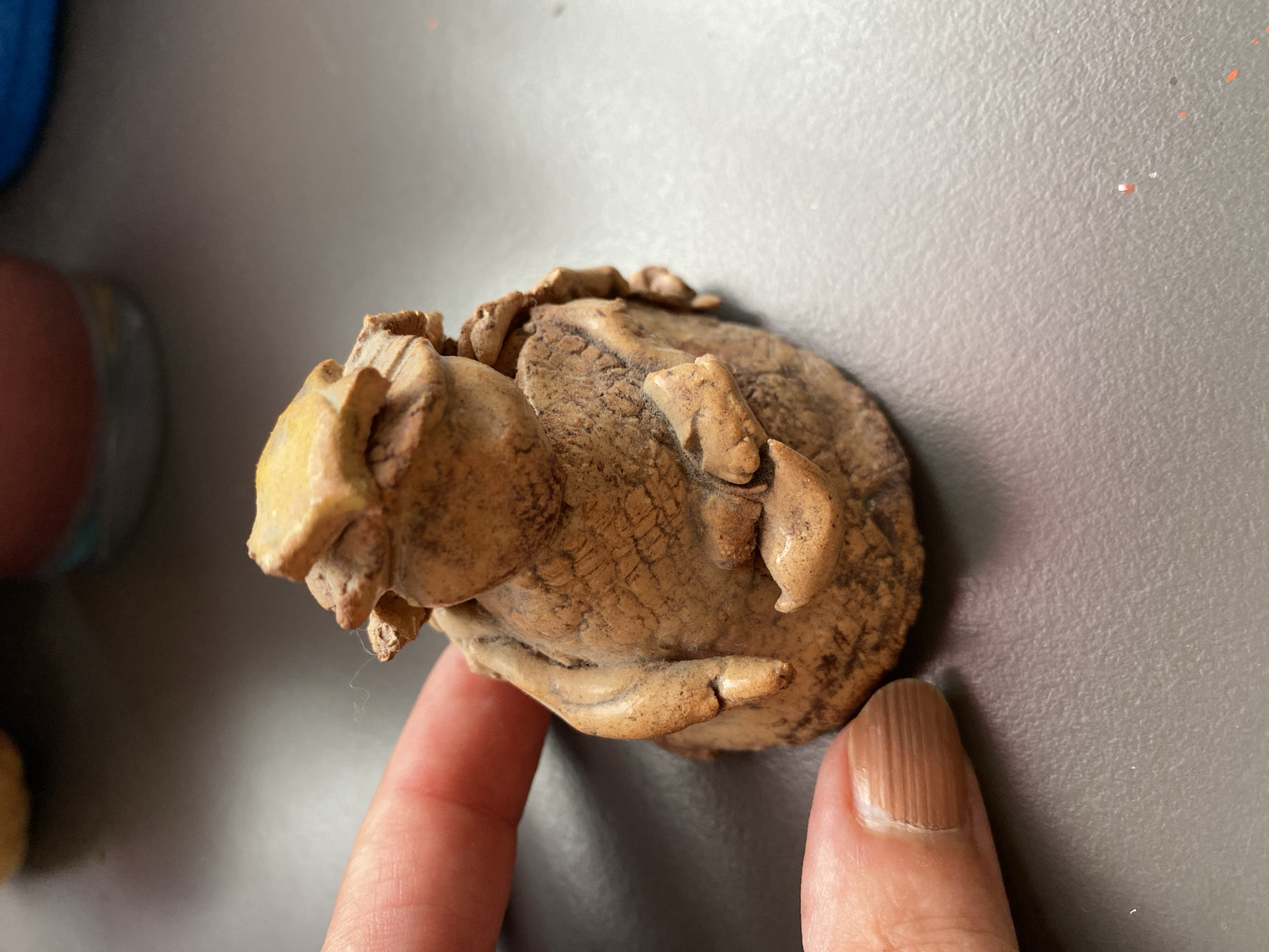 A close-up, eye-level shot shows a small, carved wooden figurine resembling a seated creature or person with a textured, layered headpiece, possibly representing a hat or headdress. The figurine is a light brown color with darker, textured lines and appears to be made from natural materials, possibly wood or a similar organic substance. The figurine's hands are visible, positioned in front of its body. A person's thumb and index finger, with visible fingernail striations, are positioned to the left of the figurine, providing a sense of scale and indicating interaction with the object. The background is a dark gray, textured surface, possibly a tabletop or desk, with some blurry, colorful objects in the upper background. The image was captured on July 30, 2023, in Bílina, Czechia, and is a detailed close-up of the figurine and the adjacent human fingers.