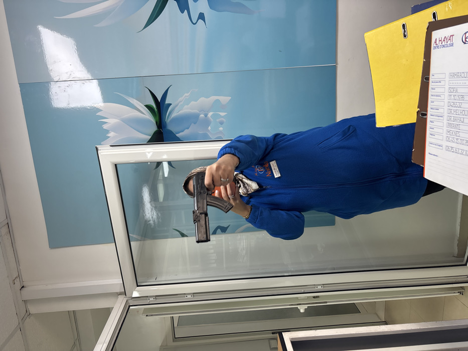 An indoor scene in a dental clinic located in Meknes, Morocco, displays a person aiming a toy or replica firearm directly towards the viewer. The individual, identified by a name tag as "Nabila Médecin Dentiste" from "Al Hayat" clinic in "Meknes," is a female. She is wearing a blue, long-sleeved, fleece-like zipped top, black pants, and a patterned headscarf that obscures her hair and part of her face. She holds a dark-colored object resembling a pistol with an exaggerated, extended rifle-style magazine (appearing similar to an AK-47 magazine) with both hands, extended forward at chest height. Her face is fully obscured by the object.
To the left, a white-framed glass door or partition is partially open, revealing a white-walled corridor. The back wall is decorated with multiple large panels featuring a light blue background and a white lotus flower design, reflected in simulated water. The ceiling is white. In the foreground on the right, a desk surface is visible, holding a yellow binder and a brown binder. A document on the brown binder shows columns of names and numbers, with "A.HAYAT DENTIST" printed at the top right, confirming the location as a dental practice. The environment is well-lit, likely by artificial overhead lighting.