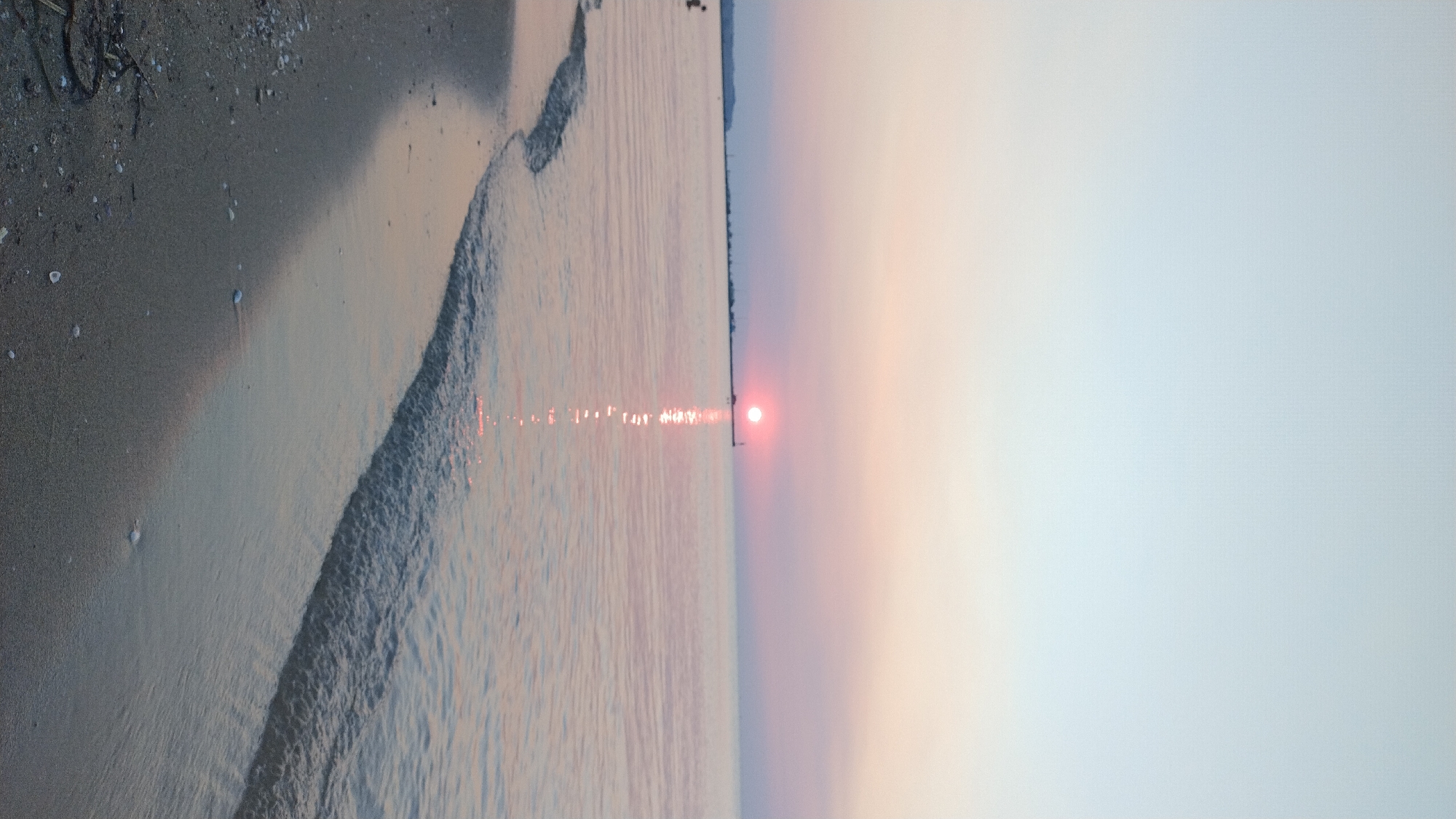 A serene sunset casts a soft pink and pastel glow over the tranquil sea at Neoi Epivates, Greece. The sun, a small, bright orb, sits low on the horizon, its reflection creating a shimmering path across the water. A long, thin pier stretches out into the sea, punctuated by distant structures and a lighthouse. Gentle waves lap at the sandy shore in the foreground, leaving behind scattered seashells and debris. The sky transitions from a pale blue at the top to a soft peach and pink hue near the horizon. A lone figure can be seen in the water to the far left, partially submerged.