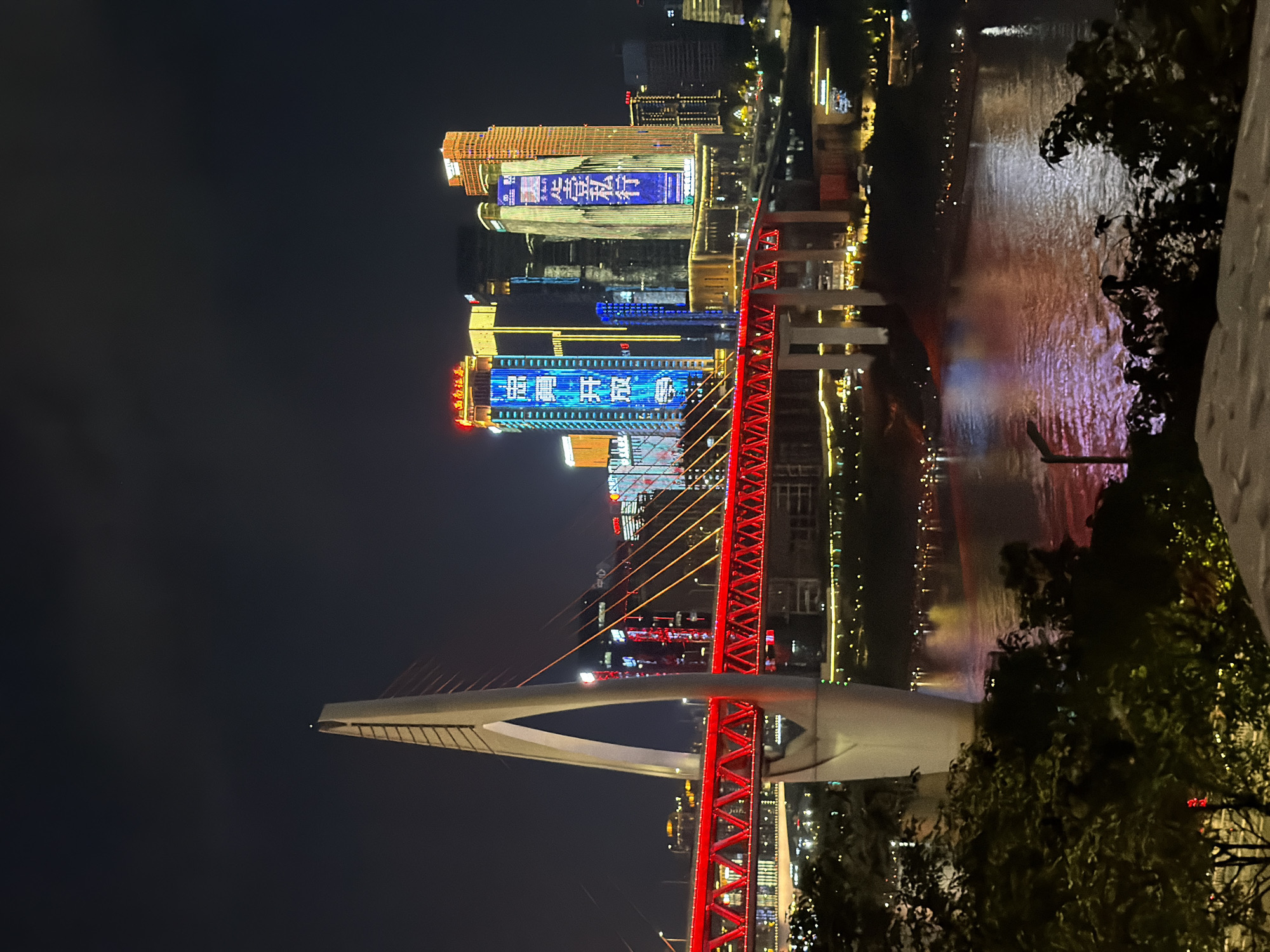 The image captures a nighttime view of a modern city skyline in Yuzhong District, China, characterized by a brightly illuminated suspension bridge and towering skyscrapers. The bridge, a prominent feature, is painted a vibrant red and spans across a body of water, reflecting the city lights. Its white pylon, a striking architectural element, rises majestically against the dark sky.
Several skyscrapers dominate the background, their facades adorned with colorful LED displays showcasing Chinese characters and advertisements. These buildings are a testament to the city's development and bustling urban life. The river below, reflecting the surrounding lights, adds to the dynamic and atmospheric quality of the scene.
The foreground features lush green foliage, partially obscuring the view and adding a touch of nature to the urban landscape. The overall impression is one of a vibrant and energetic city, alive with light and activity at night. There are no visible people in the image.
