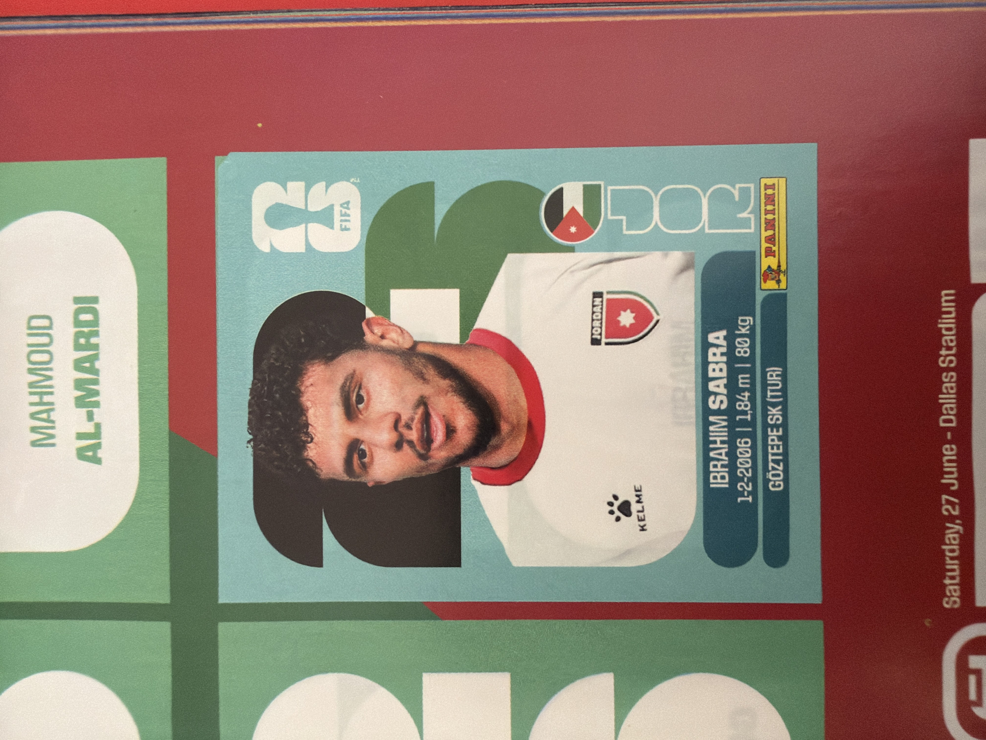 The image is a close-up of a soccer sticker album. The primary subject is a sticker of a male soccer player with curly dark hair and a beard. He is wearing a white jersey with red trim and a badge on the left chest that says "JORDAN" and features a star. The player's name, "IBRAHIM SABRA," is printed below his image, along with his birthdate "1-2-2006," height "1,84 m," weight "80 kg," and his club "GÖZTEPE SK (TUR)". A "PANINI" logo is visible in the bottom right corner of the sticker. The background of the album page is a light blue, with a large black numeral "2" partially visible behind the player. To the left of the player's sticker are portions of other green and white graphic elements of the album. Above the main sticker, the words "MAHMOUD AL-MARDI" are visible in green text on a light background. The album appears to be placed on a red surface, possibly a table or another book. The image does not provide any information or context about the city of Zlatitrap, Bulgaria.