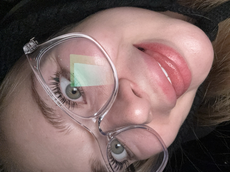 A close-up, rotated perspective of a person wearing transparent glasses. The glasses reflect a rectangular green and yellow light. The person has light skin, full lips with shiny pink lipstick, and grey-green eyes with dark lashes. A black fabric covers the top of their head.