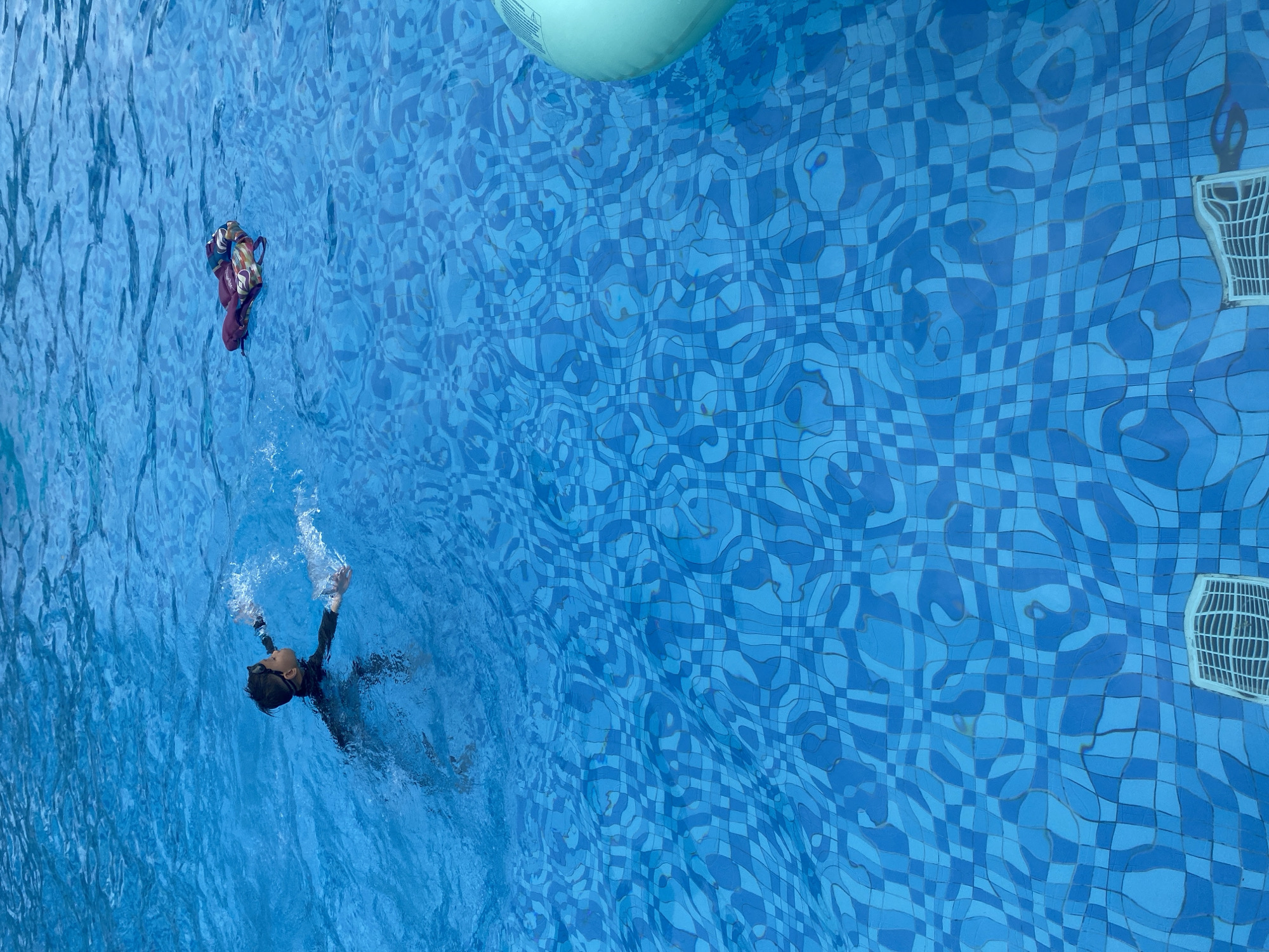 An overhead shot shows a person swimming in a pool with light blue water and tiled bottom, in Cam Lam, Vietnam. A partially submerged inflatable floats at the top of the frame and a pool filter is partially visible at the right. Another inflatable can be seen in the top left corner of the image. The swimmer is wearing dark clothing and goggles and facing toward the left of the frame.