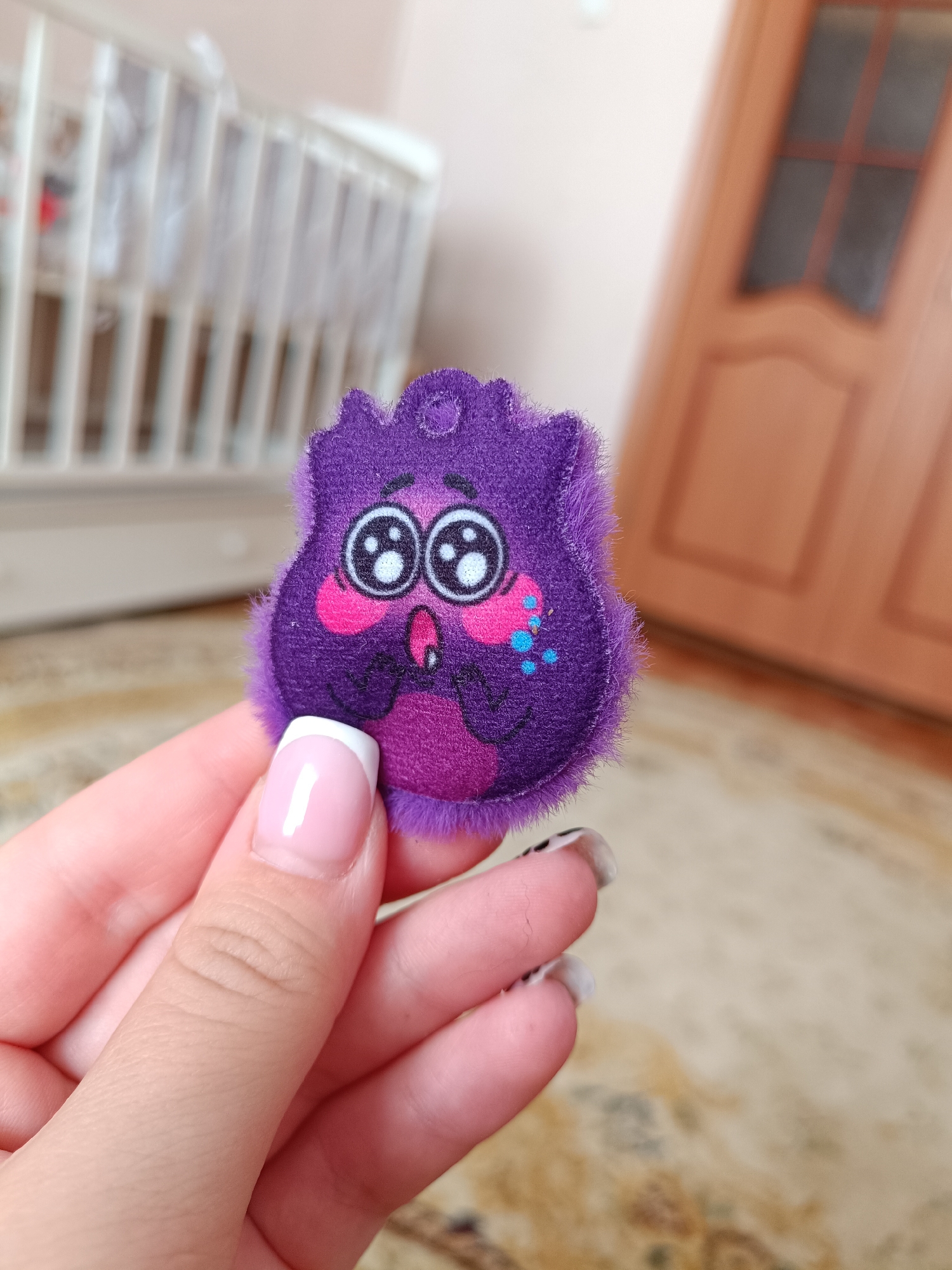 A medium shot depicts a hand holding a purple, cartoonish, fuzzy, felt creature with wide eyes and pink cheeks. The fingernails on the hand are manicured, some with French tips and others with a black and white pattern. In the background, out of focus, is a white crib with vertical slats, a beige carpet, and a brown wooden door. The location in the city of Tobolsk, Russia is not discernible.