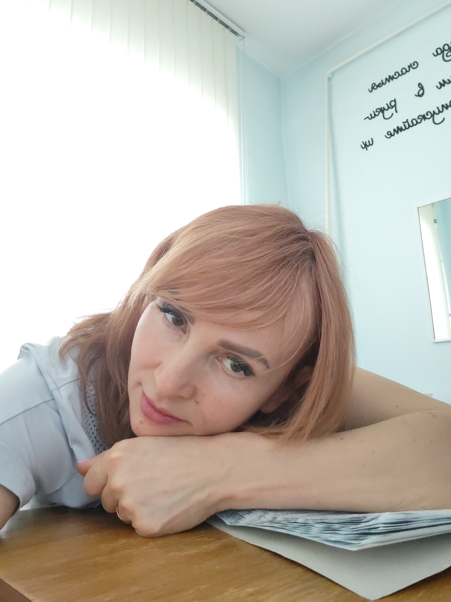 This image depicts an adult female in an indoor setting within the city of Chaikovsky, Russia. The subject, with shoulder-length, light reddish-brown hair and bangs, is positioned towards the left of the frame, resting her chin on her left hand, which in turn is placed on her right forearm. Both arms rest on a stack of white papers, possibly banknotes, displaying blue and green markings, situated on a light brown wooden table. A gold-colored ring is visible on the ring finger of her left hand. She wears a light blue short-sleeved garment, possibly a polo shirt or scrub top, with a visible mesh detail on the shoulder. Her face is lightly made up, featuring mascara and shaped eyebrows, and she exhibits a neutral to slightly smiling expression. The background consists of a light blue wall. On the right, a white panel on the wall displays black, mirrored Cyrillic text, which is likely a reflection. Below this text, a portion of a rectangular mirror with a simple frame is visible. The upper left portion of the image is dominated by an overexposed white window fitted with vertical blinds, indicating a bright light source.