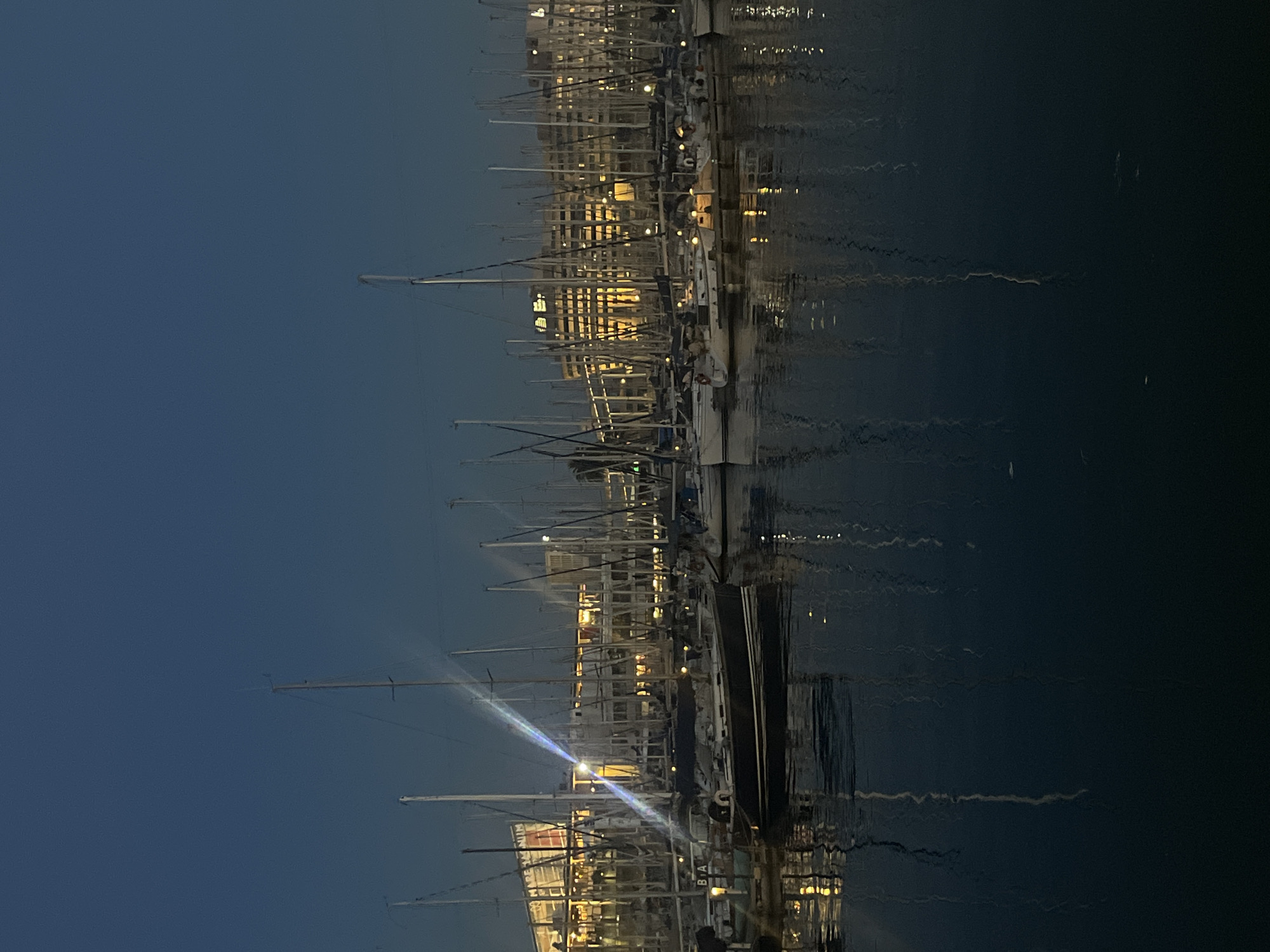 The image captures a marina in Barcelona, Spain, at dusk or night. A line of sailboats is moored at the dock, their masts pointing upwards towards a dark blue sky. The city lights and the reflections on the water create a shimmering effect. The buildings of the city appear in the background, their lights contrasting with the darkening sky.