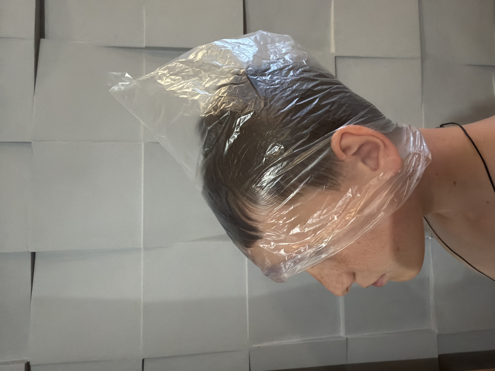 The image shows a close-up profile view of a young person with a clear plastic bag covering their head, obscuring their hair and part of their face. The person is wearing a black string around their neck. The background consists of a wall with a textured, grid-like pattern, appearing to be made of rectangular panels. The lighting is soft and even, casting no harsh shadows. There is no visible indication of the location in Yekaterinburg, Russia, or any specific environmental context beyond the interior wall. The scene is static and appears to be a still photograph.