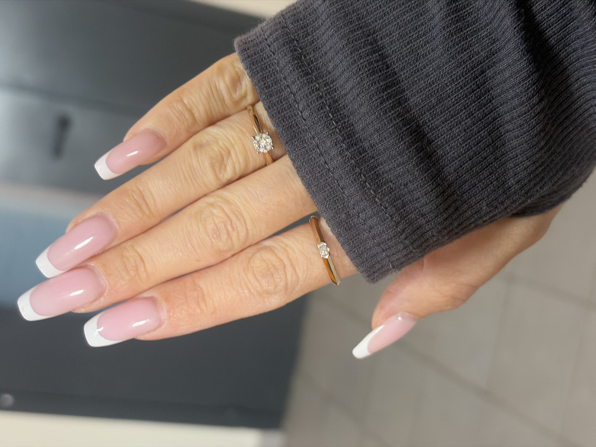 A close-up shot depicts a person's left hand wearing a gray sleeve. The hand is adorned with two gold rings, one on the ring finger and another on the pinky finger, each appearing to feature a small gemstone. The nails are styled with a French manicure, featuring a pale pink base and white tips. The background includes a dark gray or black surface to the left and a light gray or white surface to the right. The scene does not depict the city of Saint Petersburg, Russia.