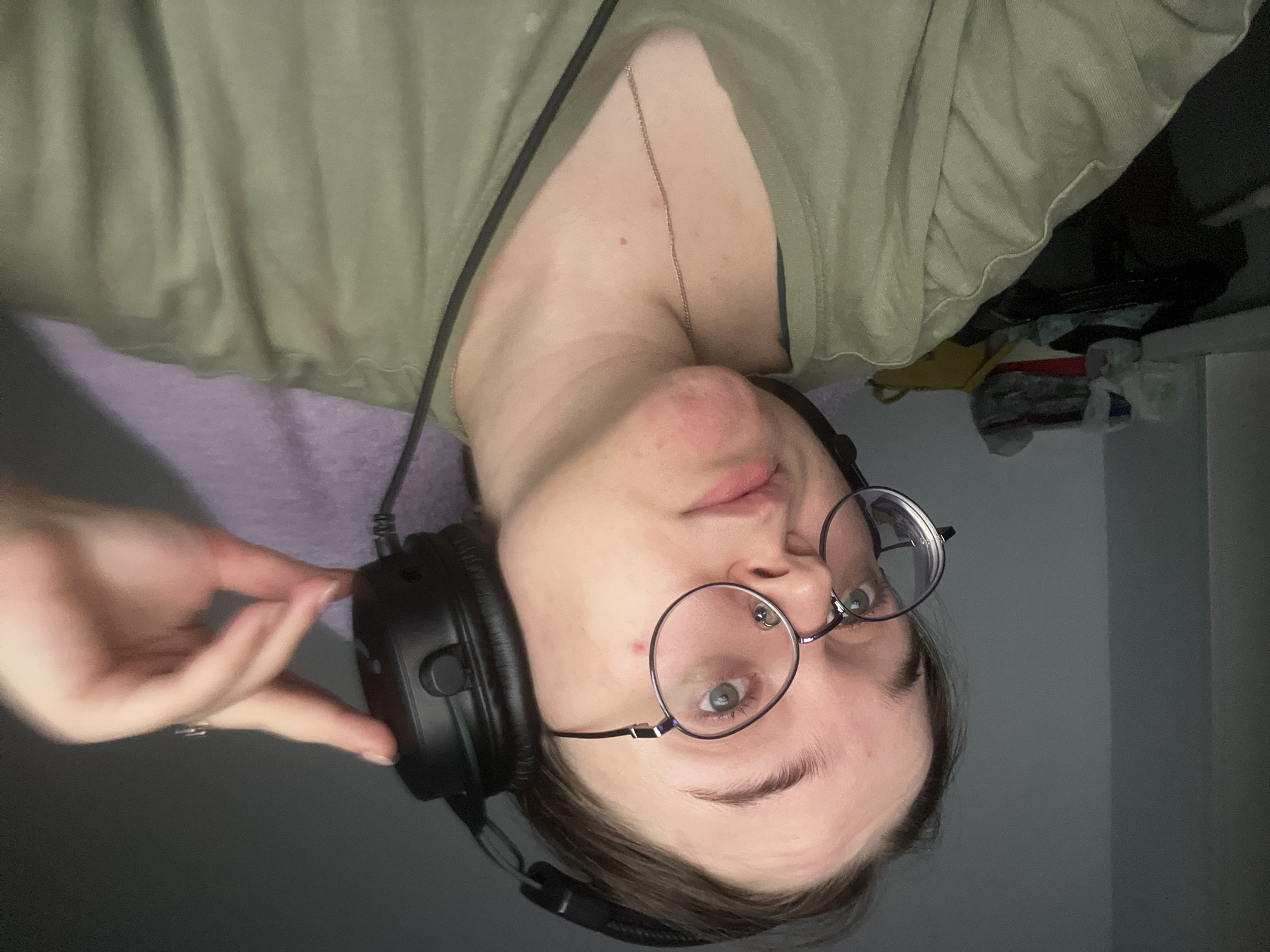 A person with fair skin, wearing round glasses, a green shirt, and black headphones, is seen in a portrait-style image. The person has green eyes and a nose piercing. Their left hand is visible, adjusting the headphones. A silver necklace adorns their neck. The background includes a gray wall and indistinct items suggestive of a bedroom or personal space. There is no evidence to suggest that the image was taken in Pushchino, Russia.
