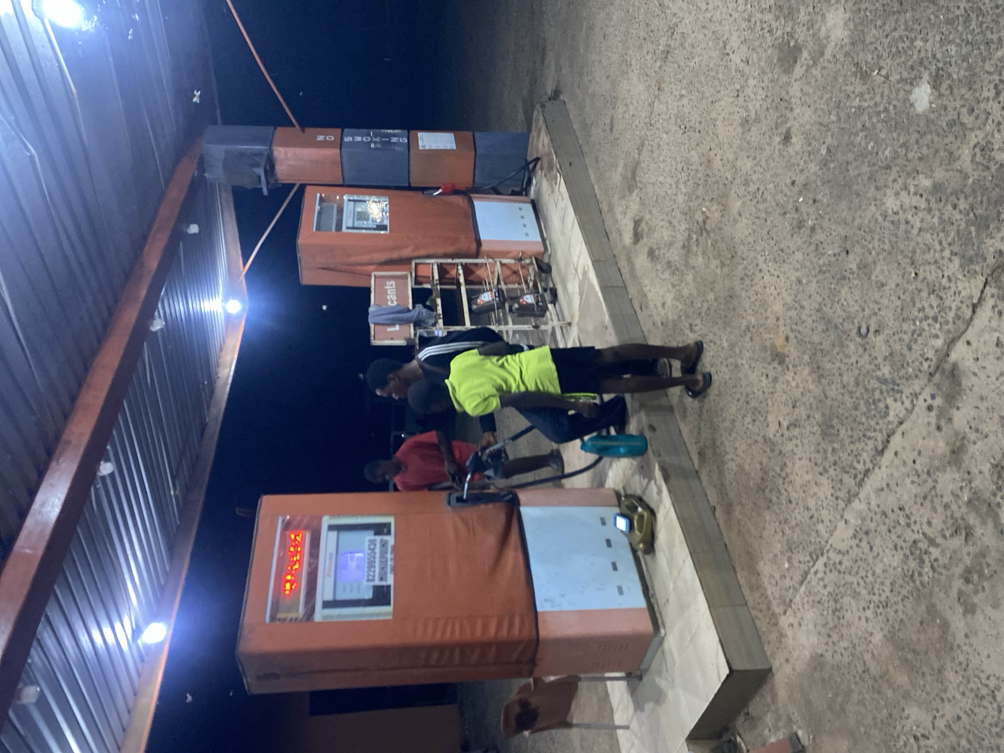 The scene depicts a nighttime view of a fuel station located in Sasaro, Nigeria. The area is illuminated by several bright, exposed light bulbs affixed to the underside of a corrugated metal roof structure, supported by orange metal beams. The ground surface is a coarse aggregate concrete.
In the foreground, there are two fuel dispensers. The left dispenser is orange on its upper section and light blue on its lower half. Its digital display shows red characters and a lower panel with "8229955438" and "HOMEPOINT" text. A fuel nozzle from this dispenser is actively in use. A yellow plastic jerrycan and a blue plastic bottle are placed on the ground near its base. A brown plastic chair is positioned to the left of this dispenser. Three males are present. One male, wearing a red t-shirt, stands behind the left dispenser. Another male, in a bright green t-shirt and dark shorts, stands in front of the left dispenser, facing right, with his hands near the engaged fuel nozzle. A third male, wearing a dark top with white stripes, stands to the right of the male in green, adjacent to a metal utility cart.
The right fuel dispenser is also orange and light blue, and its nozzle is holstered. A metal utility cart with shelves, holding several small containers (likely lubricants) and draped with a grey cloth, is positioned between the right dispenser and a dark grey and orange pillar. This pillar displays "NO SMOKING" in vertical white text on its dark grey section. The background beyond the illuminated canopy is dark, indicating night.