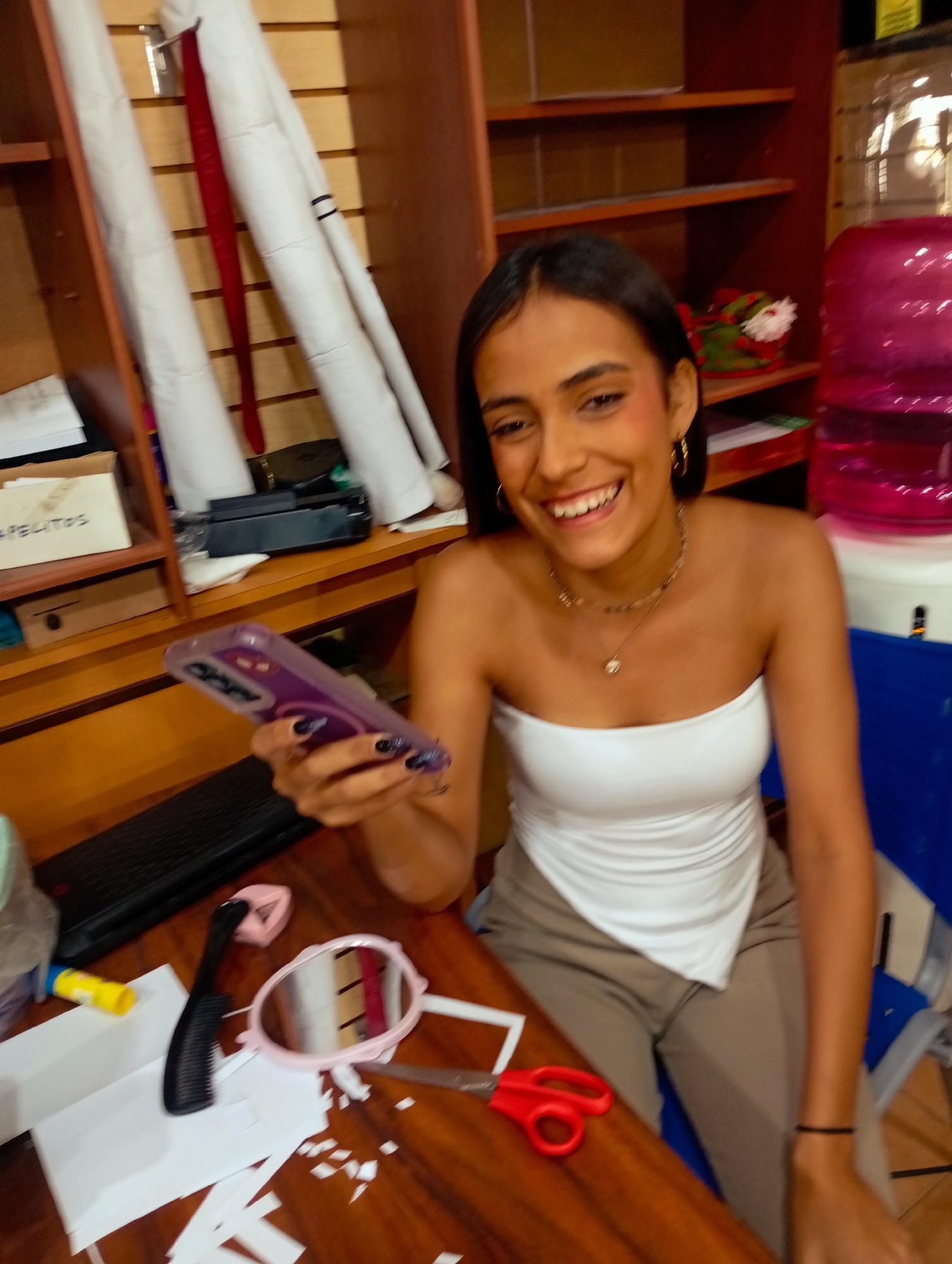 A smiling woman is indoors, holding a smartphone with a purple case. She is wearing a white strapless top and light brown pants. On the wooden table in front of her are scissors, a comb, a small mirror, and paper scraps. Behind her is a shelf with various items, including rolled-up white fabrics and a water cooler with a pink bottle. The location is Juan Griego, Venezuela, suggested by the style of the environment.