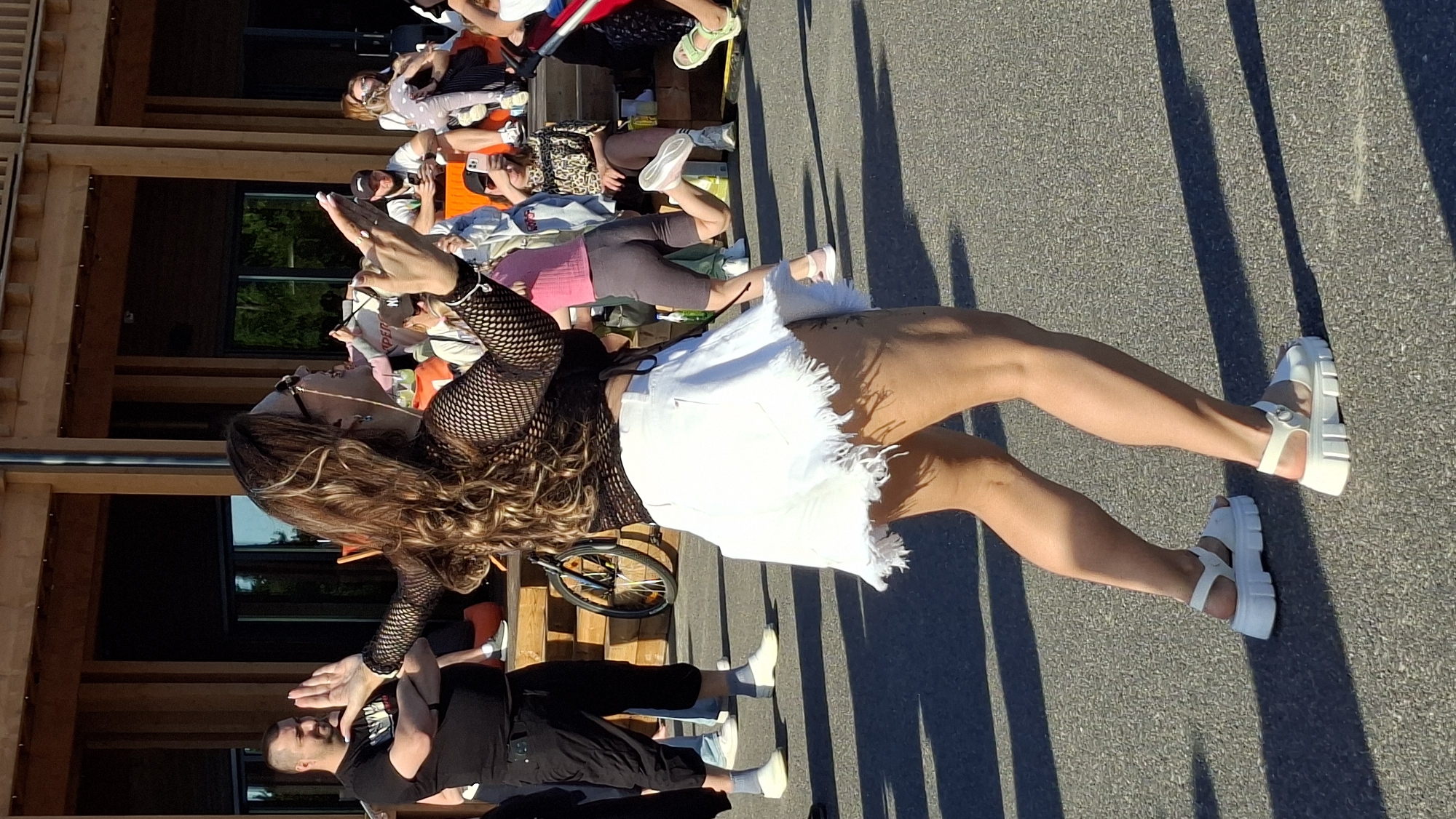 A medium shot captures a woman dancing in the foreground, her back to the camera, dressed in a black mesh top and a white frayed denim skirt. She wears large white platform sandals. Her arms are raised, and her head is turned to the right, with long, wavy brown hair cascading over her shoulders. Behind her, a crowd of people is visible. To the left, a man in a black t-shirt and black shorts stands with his arms crossed, looking towards the woman. A bicycle is parked behind him. In the background, a wooden structure with a covered area and steps suggests an outdoor venue. The ground is asphalt, with shadows cast by the sunlight. The scene is set in a public outdoor space, possibly a festival or event, in Tallinn, Estonia, given the context of previous images.