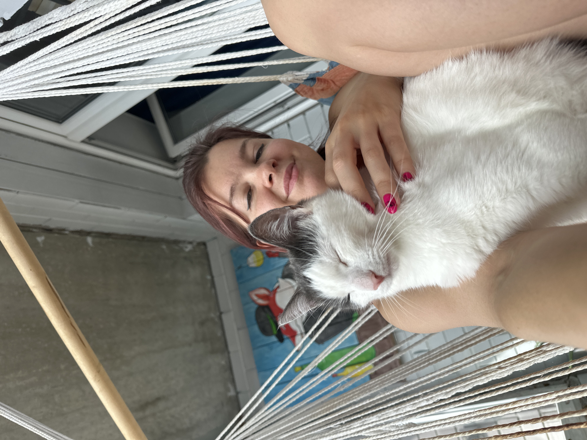 A woman is lying in a hammock holding a white and grey cat. The woman is fair-skinned with brown hair and is smiling with her eyes closed. She has pink nail polish on. The cat is mostly white with grey patches on its head and is sleeping. The hammock is made of white rope and has a wooden bar at the edge. The background shows a concrete wall and a window. There is a mural on the wall with a red fox, a black bicycle, and a green bottle. The photo was taken in Vladimir, Russia.