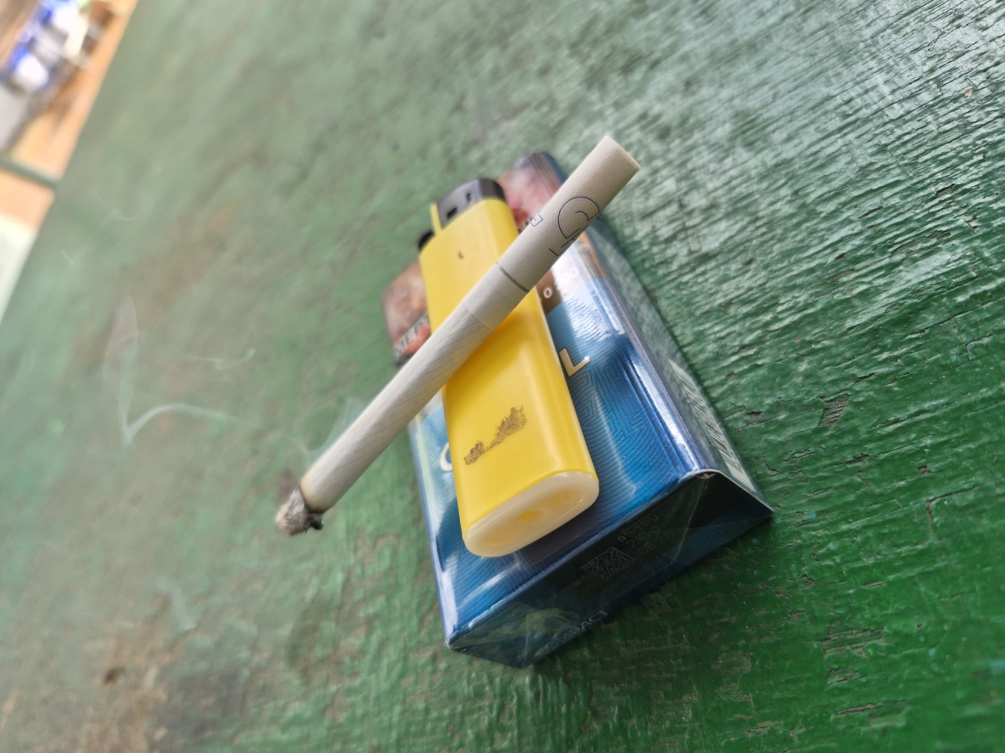 A lit cigarette is placed on top of a pack of cigarettes and a yellow lighter, resting on a weathered green wooden surface. The cigarette is emitting a wispy trail of smoke, curving upwards and to the left. The cigarette pack is blue with white and silver lettering, and the lighter is a bright yellow plastic disposable lighter. The wooden surface has a textured, worn appearance with visible grain and cracks, painted green. The background is blurred, showing hints of an outdoor setting with muted colors. No people are visible in the image. The scene is static and does not depict any ongoing action. The lighting suggests it is daytime, possibly outdoors, given the natural light and textured environment.