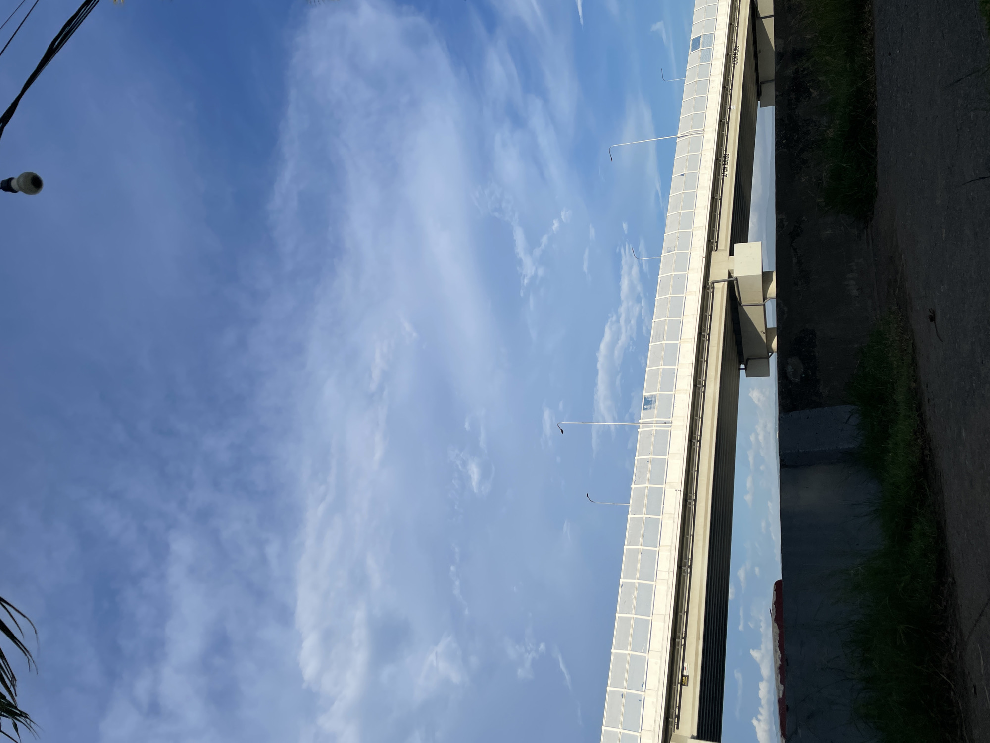 The image shows a bridge over a highway under a cloudy blue sky. The bridge has a white barrier along its side and several streetlights. Below the bridge is a concrete wall with grass growing in front of it. In the distance, a red and white object is visible, possibly a boat. The scene appears to be in Davao City, Philippines, given the presence of a bridge and highway, common infrastructure in the urban landscape of the city.