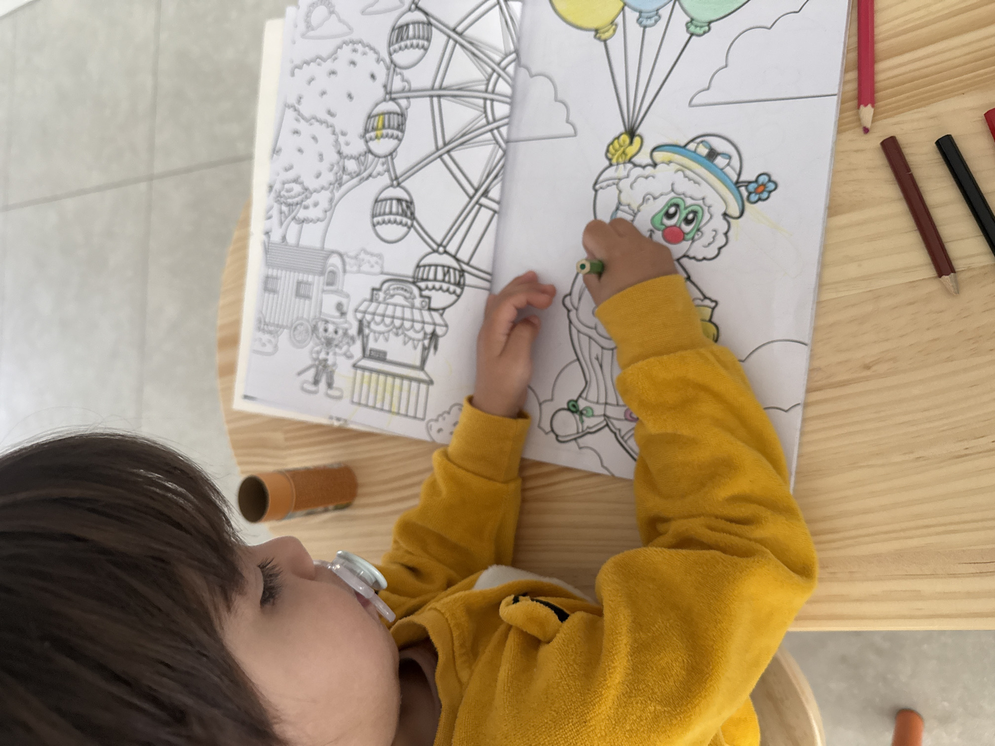 A young child with brown hair and a pacifier in their mouth, wearing a yellow sweater, is coloring in a coloring book on a light wooden table. The coloring book features a Ferris wheel and a clown holding balloons. Several colored pencils are placed beside the coloring book. There is a toilet paper roll on the table. The floor is tiled. The location is unspecified, as there is not enough information in the image to determine the location in Junglinster, Luxembourg.