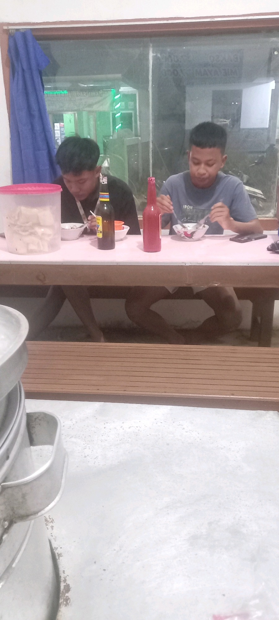 The image shows two young males sitting at a table, eating. The table is covered with a pink tablecloth, and there are two bowls of food, two bottles, and a plastic container on it. One of the males is wearing a blue t-shirt with the word "IRON" printed on it, and the other is wearing a black t-shirt. They appear to be in a restaurant or eatery in Pontang, Indonesia, judging by the background, which includes a large window showing a street scene. The overall lighting suggests it is nighttime or dusk.