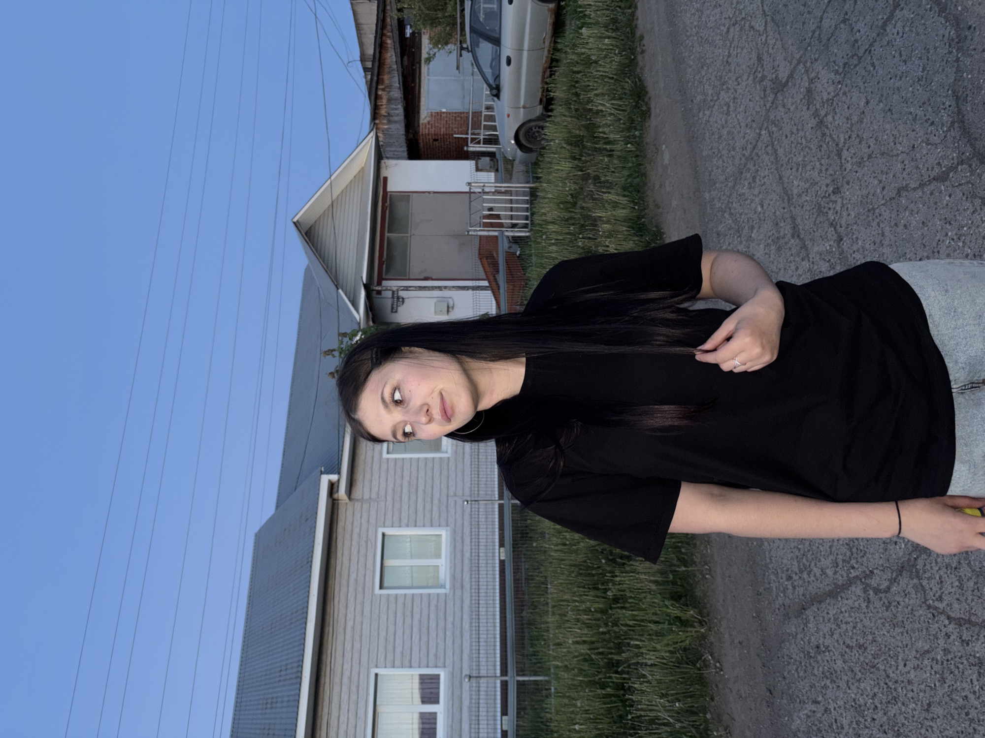 A woman is standing on a paved street in Фоки, Russia. She has long dark hair and is wearing a black t-shirt and light-wash jeans. Behind her are houses, some appearing old and worn, with a car parked near one. Utility wires are visible against a blue sky. The environment suggests a residential area.