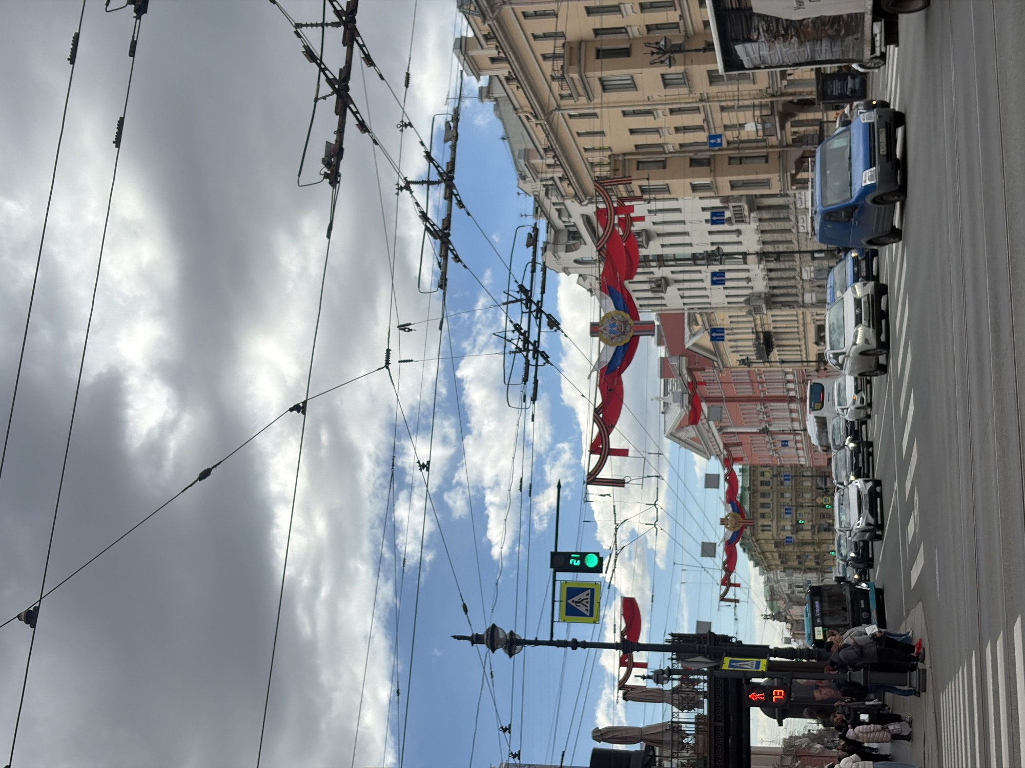 The image captures a vibrant daytime street scene in Saint Petersburg, Russia. The perspective looks down a wide city street flanked by grand, multi-story buildings of classical European architecture. On the road, a line of various vehicles, including sedans, vans (one prominent blue van), and a public bus, moves along or waits in traffic on asphalt with visible tram tracks. Pedestrians are seen on the sidewalk at the bottom right, some walking, others gathered near a streetlight.
A complex network of overhead electrical wires, likely for trams or trolleybuses, stretches across the sky, connecting to poles and buildings. The sky above is partially cloudy, showing both bright blue patches and large, grey clouds.
Festive decorations adorn the street, consisting of red and white banners strung between the buildings and wires. These banners feature a Russian-style coat of arms and the black and orange striped George Ribbons, suggesting a national holiday or patriotic celebration, possibly around Victory Day.
Visible street furniture includes traffic lights; one displays a green light with the number "21" (a countdown timer), and a pedestrian signal shows a red light with the partial text "EL". Pedestrian crossing signs and ornate streetlights are also present. The scene depicts a typical bustling day in the historic city, under a varied sky, with elements of everyday urban life mixed with celebratory decor.