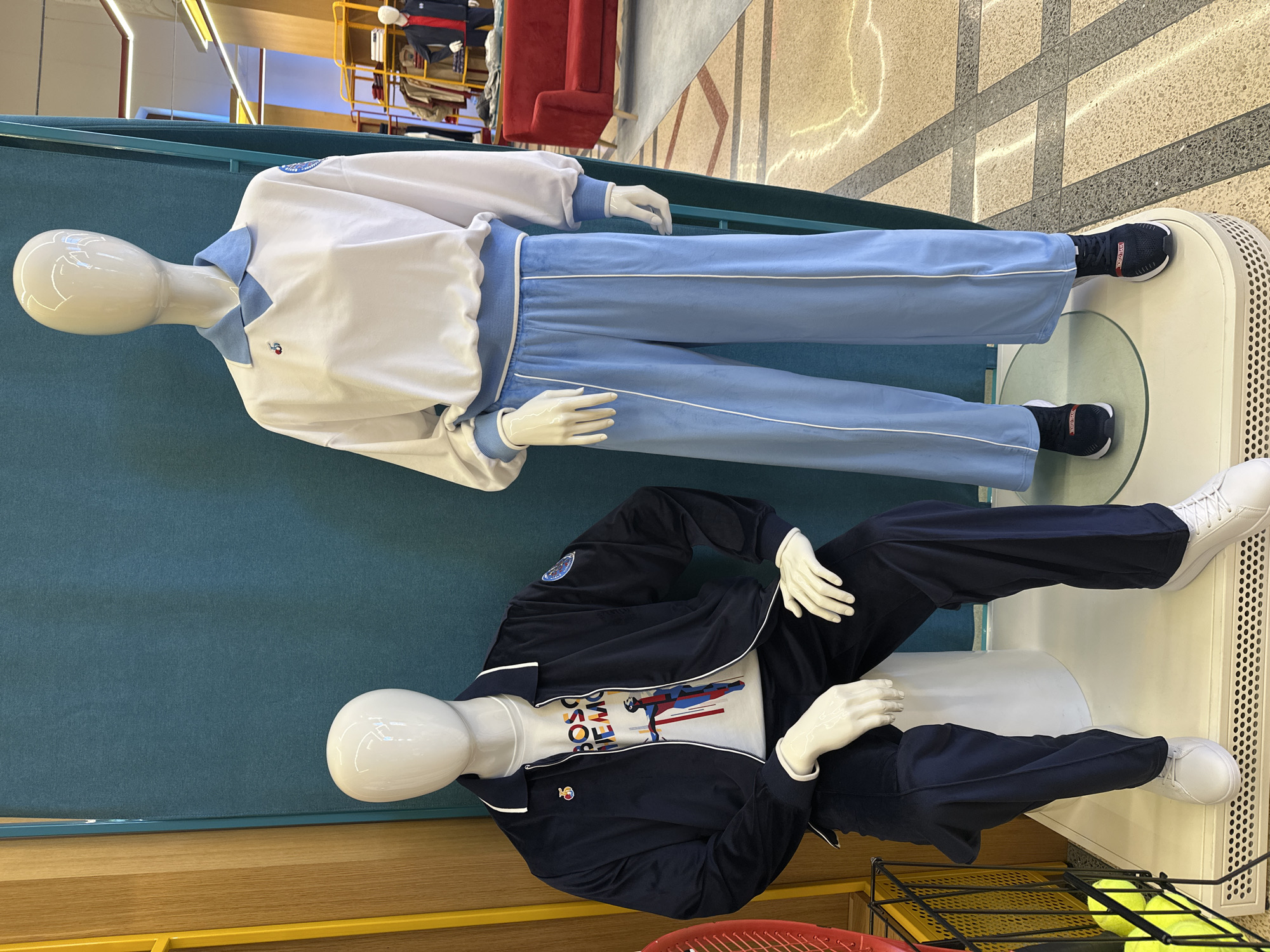 Two mannequins are displayed in an indoor setting, likely a retail store or exhibition space, with a backdrop of teal and wooden panels, and tiled flooring. The mannequins are posed horizontally, as if lying on elevated platforms.
The upper mannequin wears a white long-sleeved sweatshirt with a light blue collar and ribbed cuffs, paired with light blue track pants featuring white side stripes. Small red and black logos, possibly a stylized bird, are visible on the left chest of the sweatshirt. Its dark sneakers have partially visible text "ROC" on the side.
The lower mannequin is dressed in a navy blue zipped track jacket with white piping, worn over a white t-shirt. The t-shirt features a colorful graphic print depicting an athlete in motion, with partially visible text that appears to read "BOSS MEMO" or similar. A small logo, similar to the one on the upper mannequin, is on the jacket's left chest, and a circular blue patch is on the left sleeve. This mannequin wears navy blue track pants and white sneakers.
In the background, a red sofa and yellow metal shelving are visible. A black metal basket containing several yellow tennis balls is positioned near the lower mannequin's feet. The overall impression is a vibrant and modern display of sportswear. The context is Sochi, Russia.