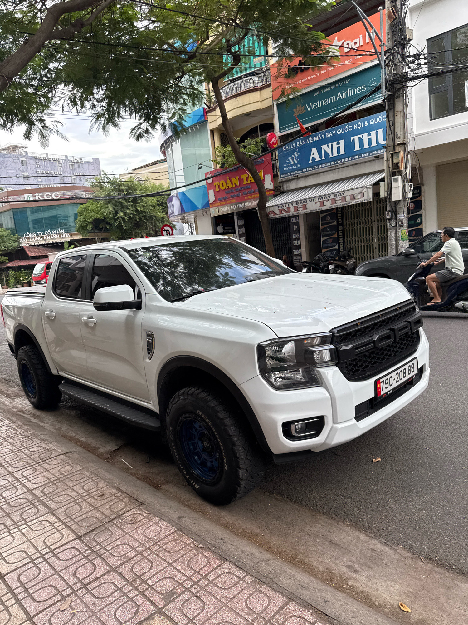 A white Ford Ranger pickup truck is parked on the side of a street in Nha Trang, Vietnam. The truck is facing left and is positioned at an angle to the curb. The license plate reads "79C-208.88". The truck has aftermarket blue wheels. In the background, there are buildings with various signs, including "Vietnam Airlines" and "Anh Thu". A man on a motorcycle is visible further down the street, moving away from the truck. The street is paved and there is a sidewalk with a patterned tile surface. Trees and overhead wires are also present in the scene.