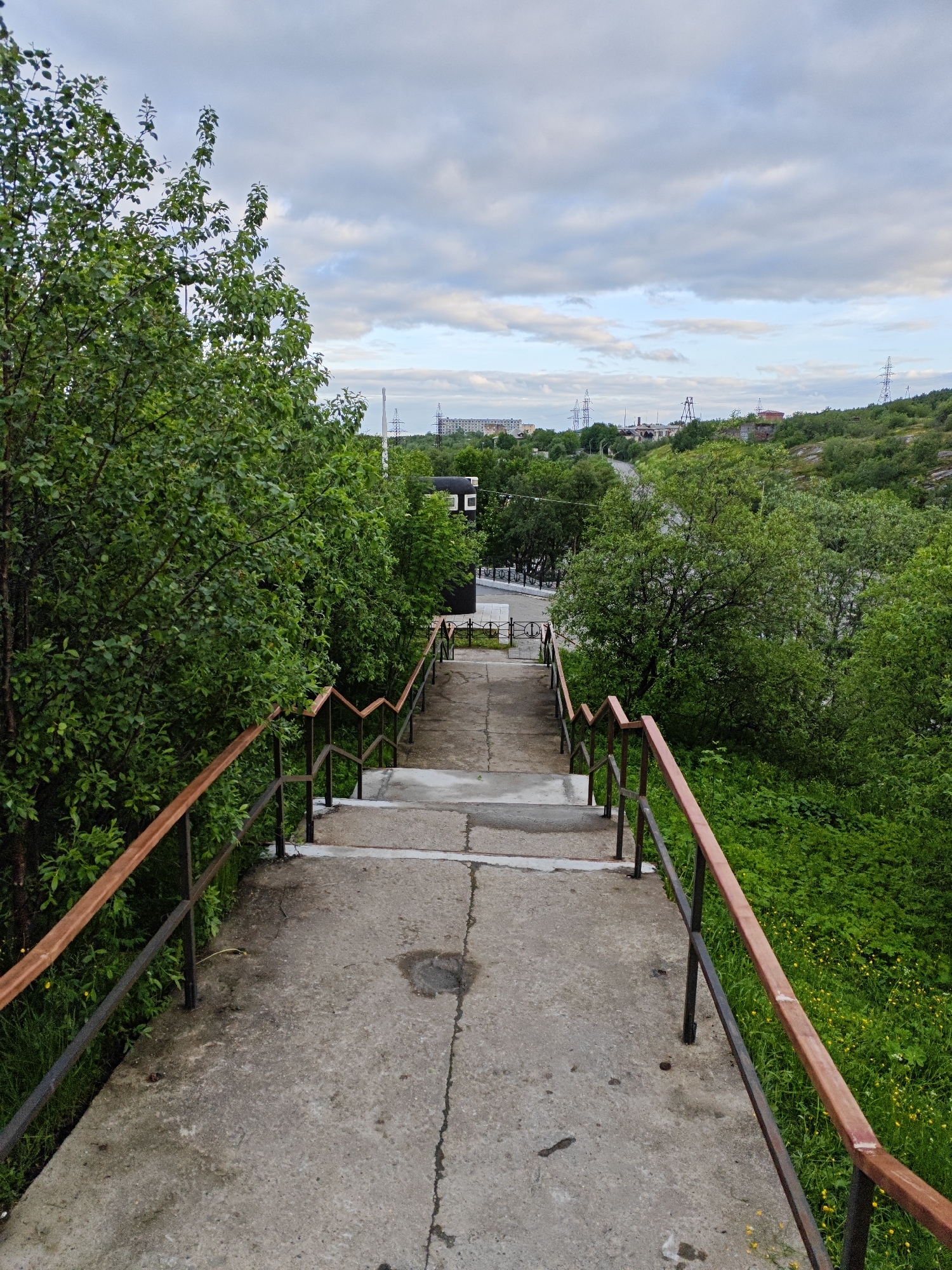 The image shows a concrete stairway leading down a gentle slope, flanked by metal railings with wooden handrails. Lush green trees and vegetation cover the sides of the stairway, with some yellow wildflowers visible in the grass. In the background, a city skyline is visible with buildings, power lines, and industrial structures, indicative of a settled, possibly industrial area. The sky is overcast with clouds. This scene is located in the city of Polyarny, Russia.
