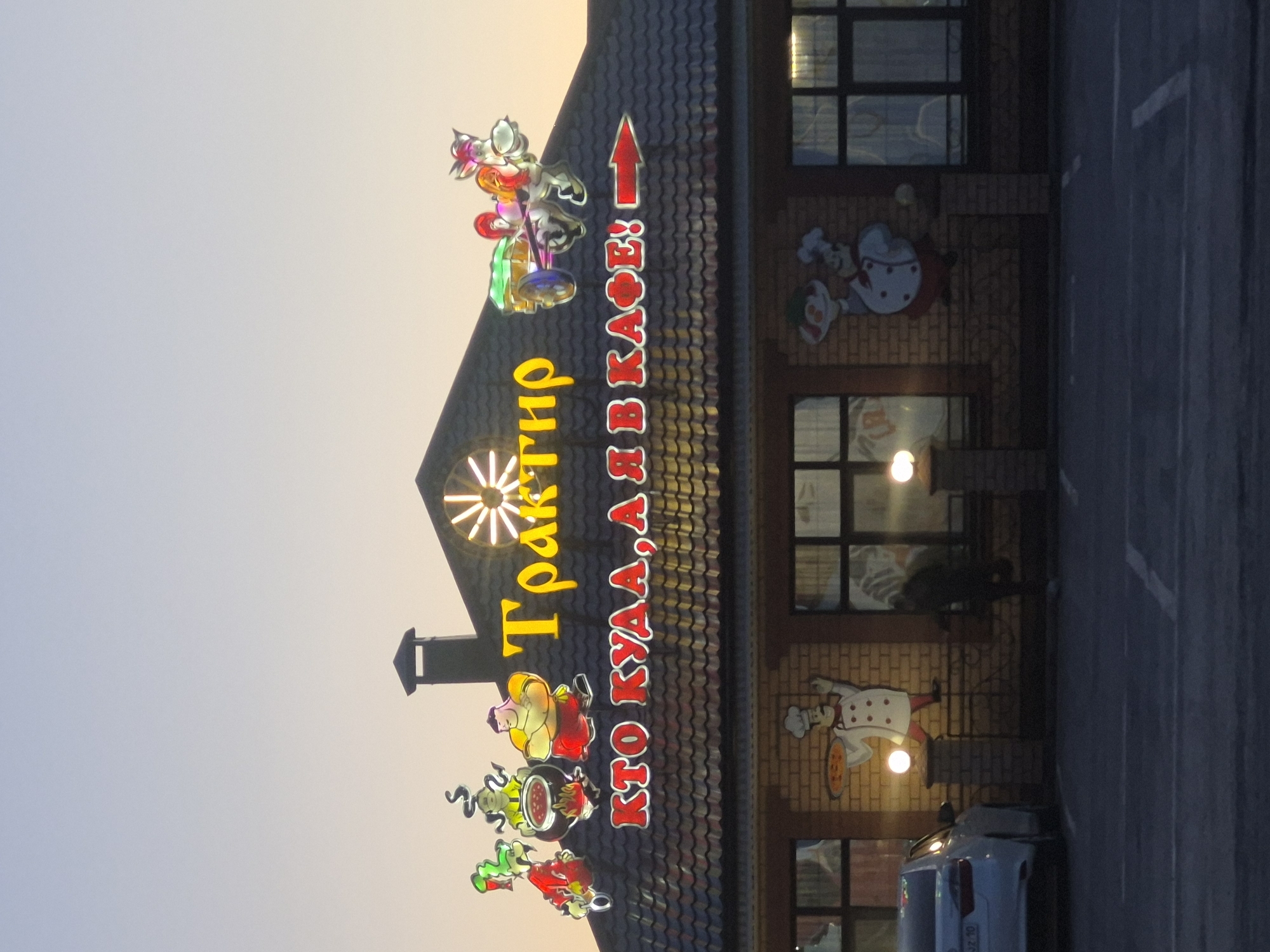 The image depicts a two-story building in Sergeevka, Russia, likely a restaurant or cafe named "Traktir." The building has a dark tiled roof with a sign that says "Traktir" in large yellow letters. Below the name, there is a red inscription in Russian, "KTO KUDA, A Y BKACE!" (roughly translates to "Who goes where, and we are in the cafe!"). Cartoonish figures of chefs and food items decorate the facade. Above the title a windmill is drawn. The building has multiple windows with dark frames, and cartoonish graphics of chefs are attached to the brick walls between the windows. A car is parked near the building on a parking lot. The sky in the background suggests either early morning or late evening.