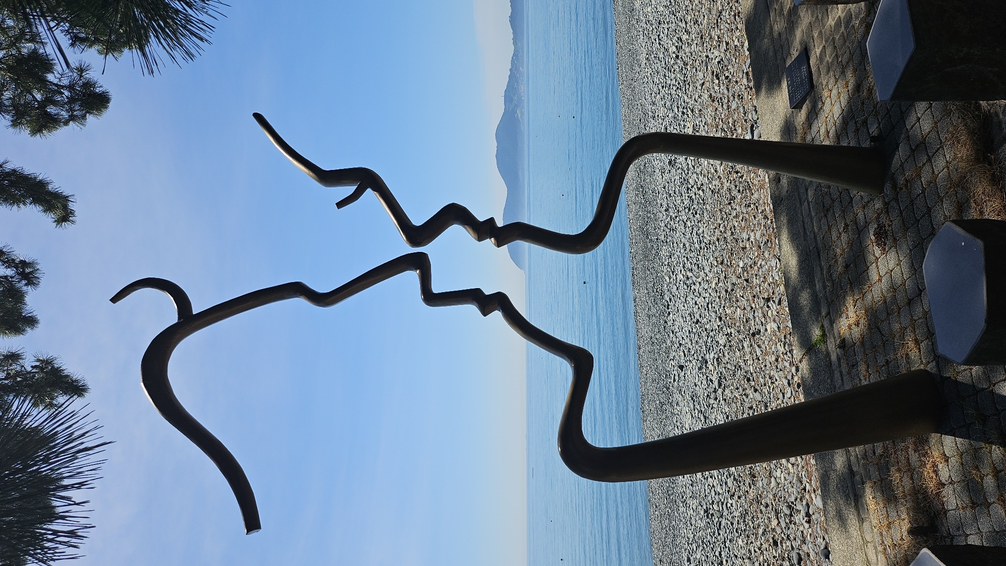 A large, abstract metal sculpture depicting two faces in profile, seemingly about to kiss, stands on a cobblestone pathway near a pebble beach. The sculpture is dark brown or bronze in color and is positioned in front of a bright blue ocean under a clear blue sky. Pine tree branches hang from the top left and top right corners of the frame, casting shadows on the sculpture and the ground. The sculpture is located in a public space, with several dark, angular benches or seating structures visible in the foreground. The sea is calm, with a distant landmass visible on the horizon. The overall scene is peaceful and serene. This sculpture is located in the city of Geoje-si, South Korea.