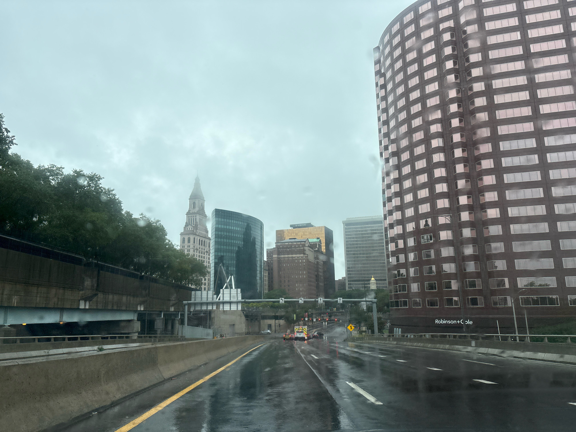 The image shows a view of a city skyline on a cloudy, wet day, likely from a moving vehicle on a highway. Several tall buildings are visible, including a distinctive brown brick building on the right with a curved facade and many windows. A tall, light-colored building with a pointed spire is visible in the distance to the left, surrounded by trees. Other modern buildings with glass facades are also present. The highway in the foreground is wet and reflective, with yellow and white lane markings. Traffic is present on the highway, including a few cars and what appears to be an emergency vehicle. A concrete barrier separates the lanes of traffic. The overall impression is one of an urban environment experiencing inclement weather. The building with the "Robinson+Cole" sign is visible on the right side of the image, indicating a business presence within the city.