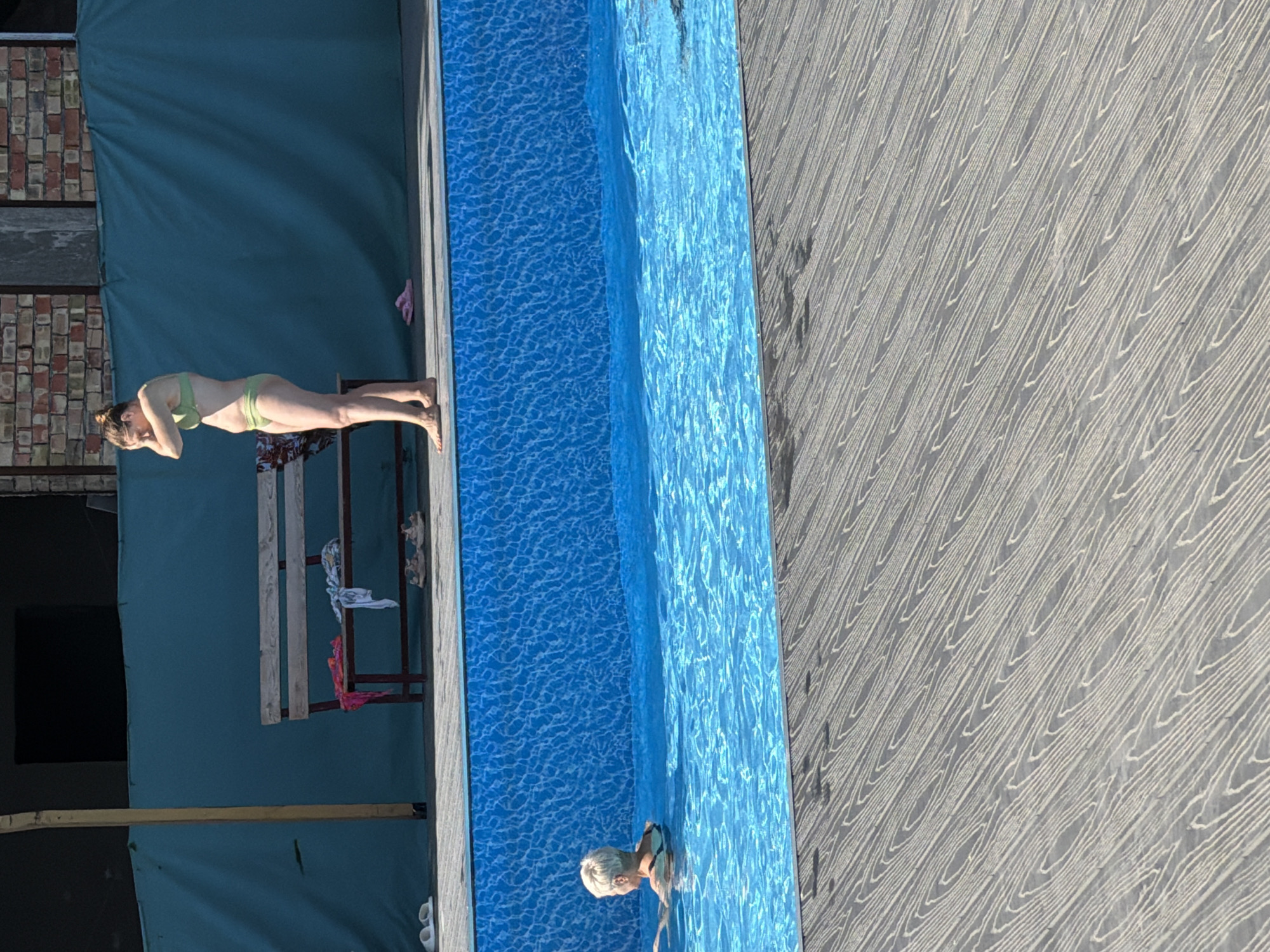 In an outdoor setting with a swimming pool, a woman in a green bikini stands on a wooden deck, touching her head. To her left, on the deck, are a bench with clothes on it and a pair of pink flip-flops. In the pool, an older woman with short white hair swims. The pool has a blue tiled interior. The deck is made of grey wood planks. In the background, a turquoise tarp hangs behind a brick structure.