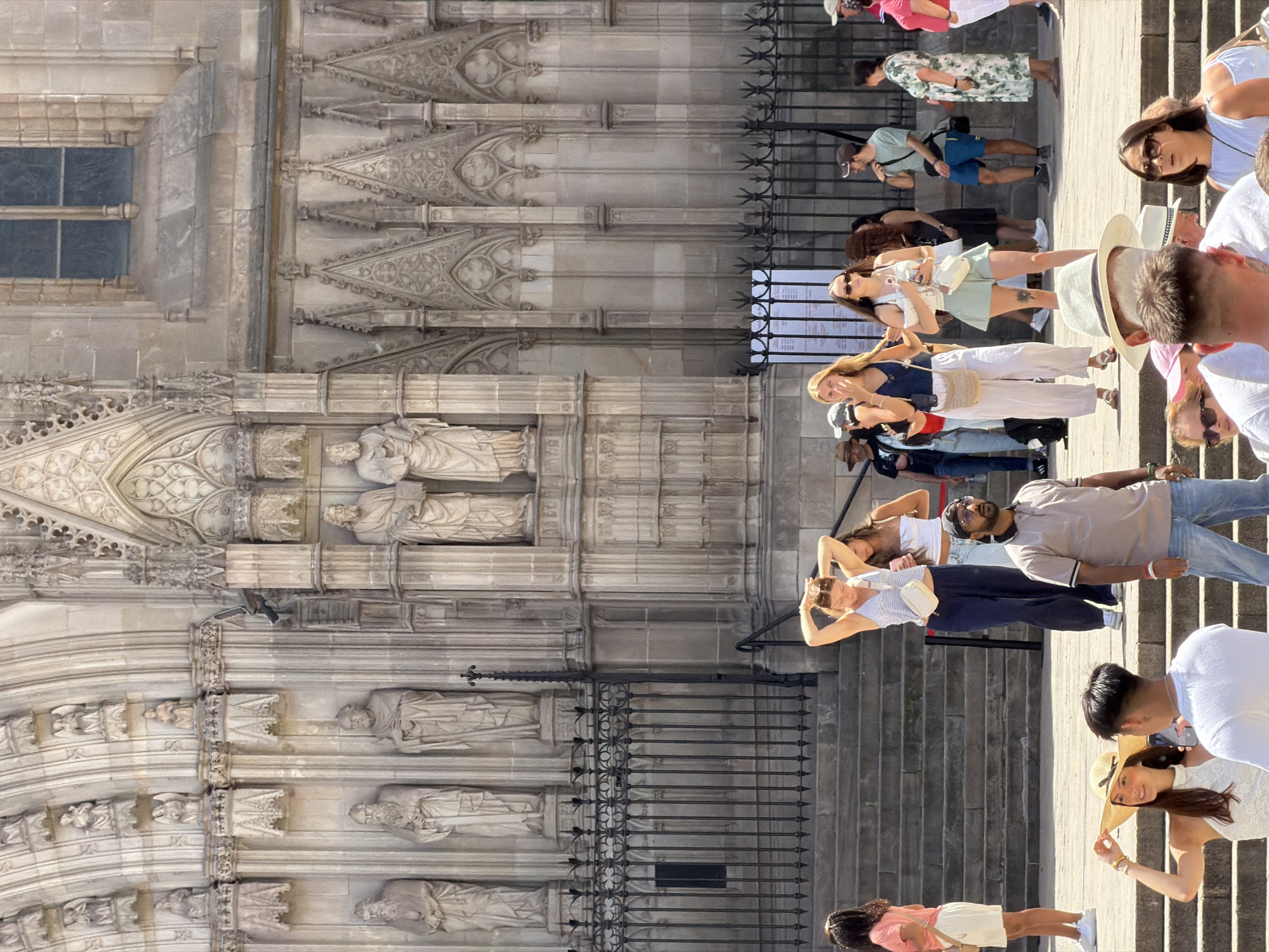 The image depicts a scene outside the Barcelona Cathedral in Spain, on a sunny day. Numerous tourists are gathered on the steps and in front of the cathedral's ornate facade. A diverse group of individuals, dressed in casual summer attire, are visible, some standing, some walking, and some engaged in conversation.
In the foreground, several people are featured. A woman wearing a wide-brimmed straw hat and a white lace top is holding the brim of her hat, looking to her right. Next to her, a man in a white t-shirt is partially visible, seemingly interacting with her. To the right of these two, a man in a grey polo shirt and jeans stands looking forward. Further back on the steps, a woman in a blue and white striped top and dark pants is adjusting her hair. Beside her, another woman in a white top and a mint green skirt is standing.
In the midground, a man in a grey t-shirt and blue shorts is seen from the side. To his right, a woman in a floral print dress is walking. Other individuals are visible further back, including a person in a pink shirt and white shorts on the far left.
The Barcelona Cathedral's imposing Gothic architecture is the backdrop, with its stone facade featuring intricate carvings and statues. A black wrought-iron fence separates the lower area from the higher steps of the cathedral. The overall atmosphere is that of a popular tourist destination with many visitors enjoying the architecture and the weather.