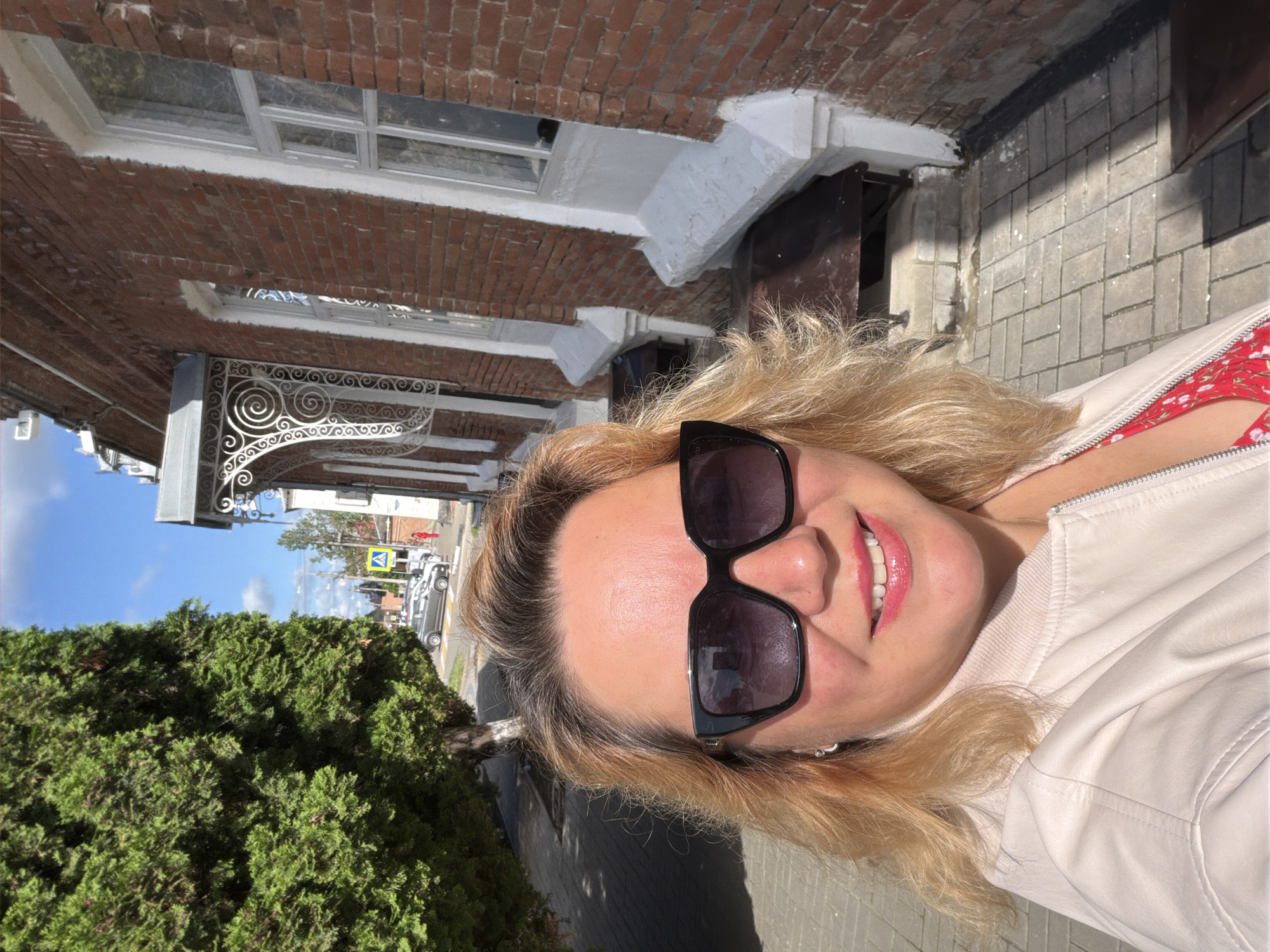 The image captures a woman outdoors, wearing sunglasses, with a building on the left and street pavement to the right. The building features a red brick facade, white window frames, and decorative ironwork. A dense green bush is visible near the bottom left. Further down the street, cars and a pedestrian crossing sign are present, and the sky is partially visible. Based on the context, the image is in Leningradskaya, Russia.
