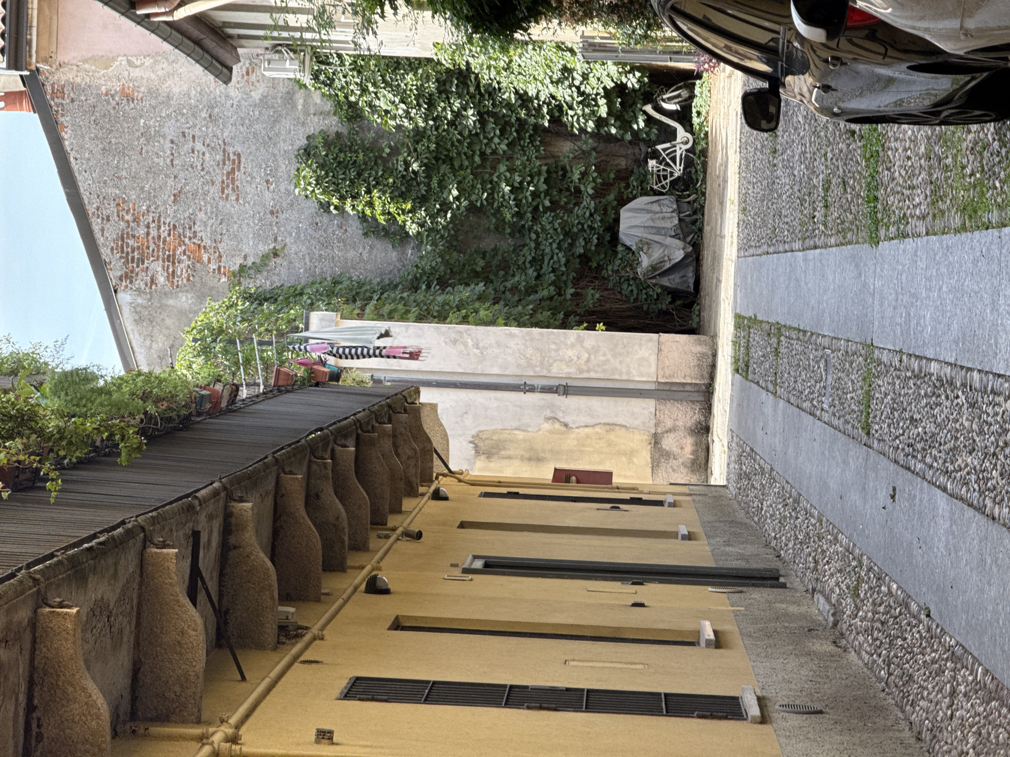 This is a ground-level, eye-level, medium shot of a narrow alleyway in Milan, Italy, on a sunny day. The left side of the alley is dominated by a yellow building with a textured, grey base. The building has several windows with black bars and a balcony with concrete supports. On the right side of the alley, a white bicycle is visible, partially covered by a grey tarp, and an air conditioning unit is mounted on a pink-toned building. The ground is paved with cobblestones and a central concrete path. In the foreground, the right side of a dark-colored car is visible. The overall scene depicts a quiet, residential alleyway in the city.