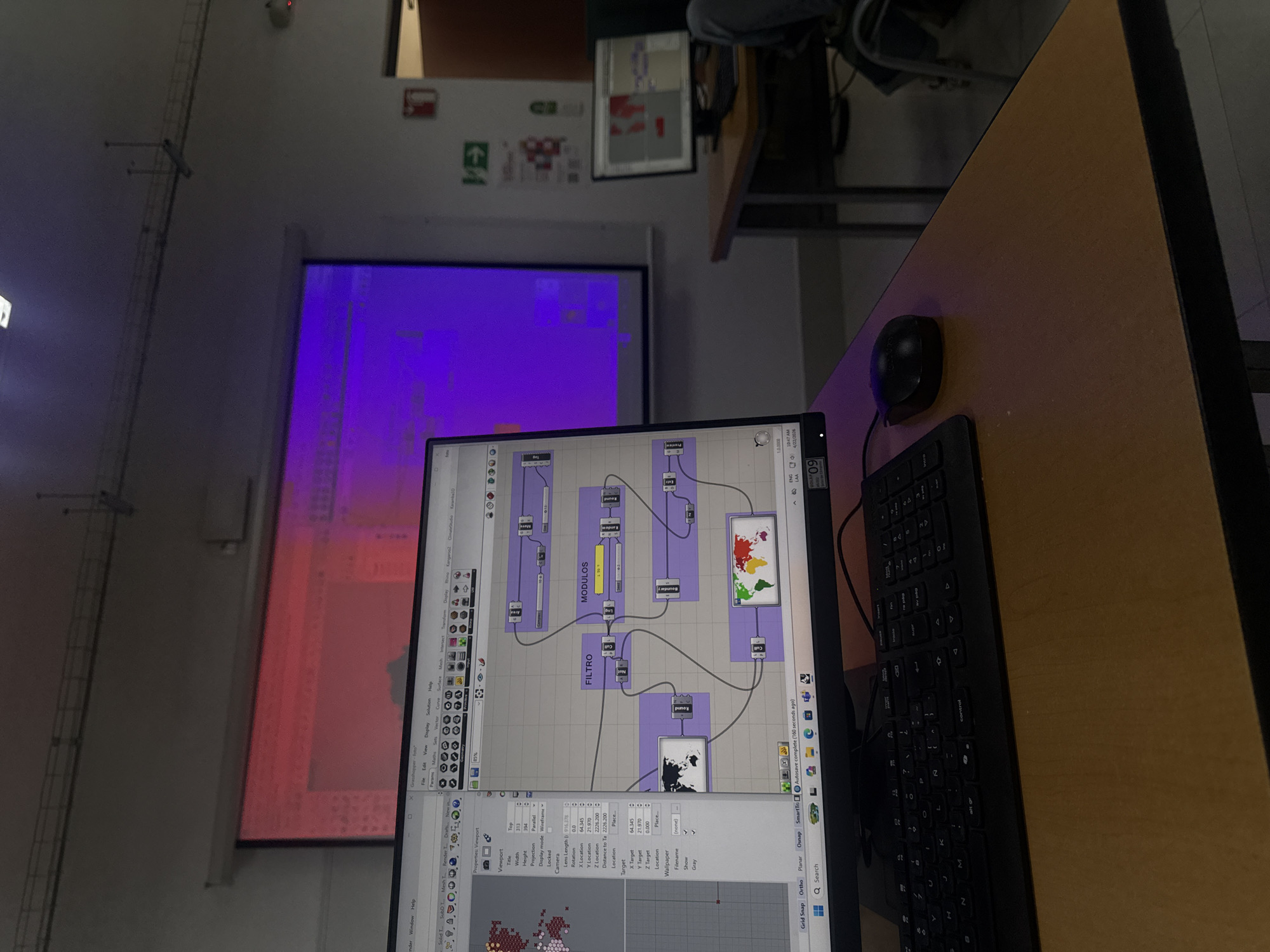 This image captures an indoor scene, likely a classroom or lab, in San Andrés Cholula, Mexico, at 10:47 AM on April 22, 2026. The main subject is a computer monitor displaying the Grasshopper visual programming environment for Rhino 3D software. The screen shows a complex node-based graph with interconnected modules, some labeled "FILTRO" and "MODULOS," alongside colorful map-like graphical outputs. A black keyboard and mouse are positioned on a light brown wooden desk next to the monitor.
In the background, a large projector screen displays a vivid, abstract pattern in magenta and red hues, which appears to be a visual output from the computer. Above the screen, an emergency exit sign with a right-pointing arrow and a fire extinguisher sign are visible on a white wall. The room's lighting is subdued, emphasizing the glow of the screens. Parts of another desk and a person's leg and arm are faintly visible in the upper background. The photo is taken from a slightly tilted, downward-looking perspective over the desk.
Visible text includes "Grasshopper," "Rhino," "FILTRO," "MODULOS," "Autosave complete (160 seconds ago)," "ENG," "10:47 AM," and "4/22/2026," along with various software interface elements and numerical data.