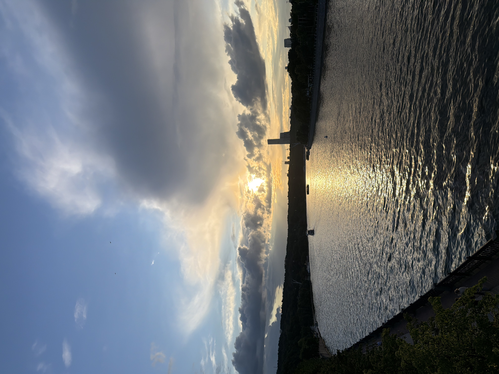This is an eye-level, medium shot of a river at sunset in an urban park. The sun is setting in the distance, casting a golden glow on the water and sky. The sky is filled with dramatic clouds, some dark and stormy, others light and wispy. The river is wide and calm, with a few boats visible in the distance. The banks of the river are lined with trees and a walking path, where a few people can be seen strolling. In the background, a few buildings and a tall, modern structure are silhouetted against the sky. The overall mood of the image is peaceful and serene, with a hint of drama from the stormy clouds.
