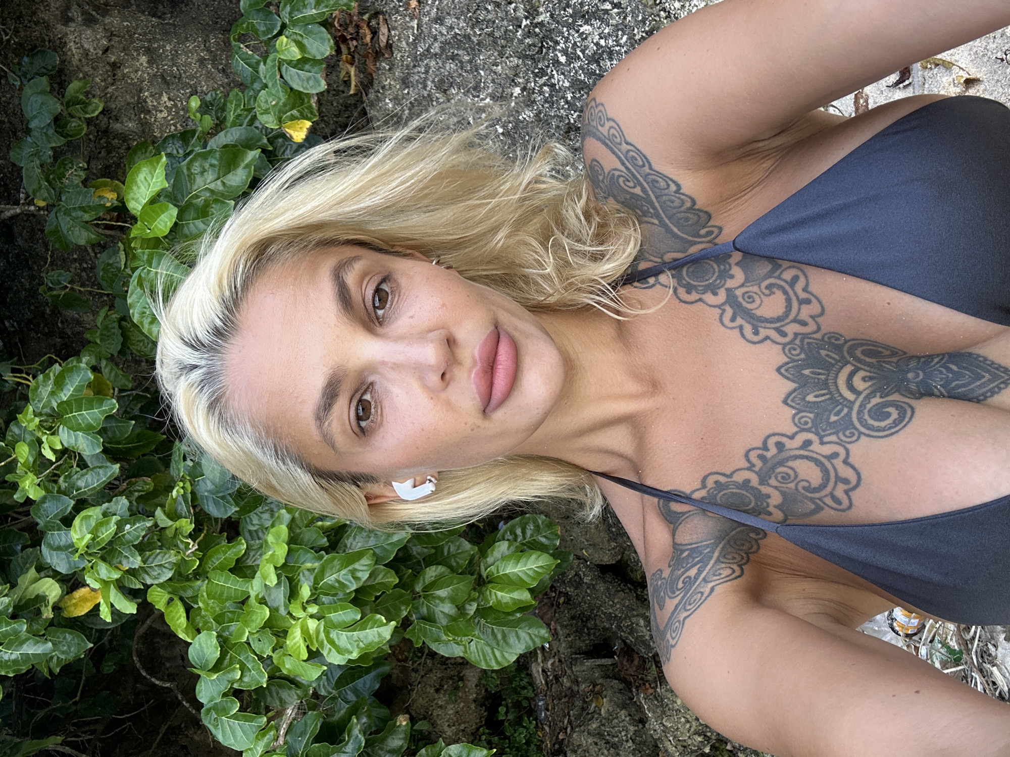 A woman with blonde hair and tattoos is pictured in a selfie. She is wearing a dark bikini top. She has a white earbud in her left ear. The background is a lush green bush and a rocky surface. The lighting suggests it is daytime. The image appears to be taken outdoors, likely in a tropical environment. The location is stated as Kuta Selatan, Indonesia.