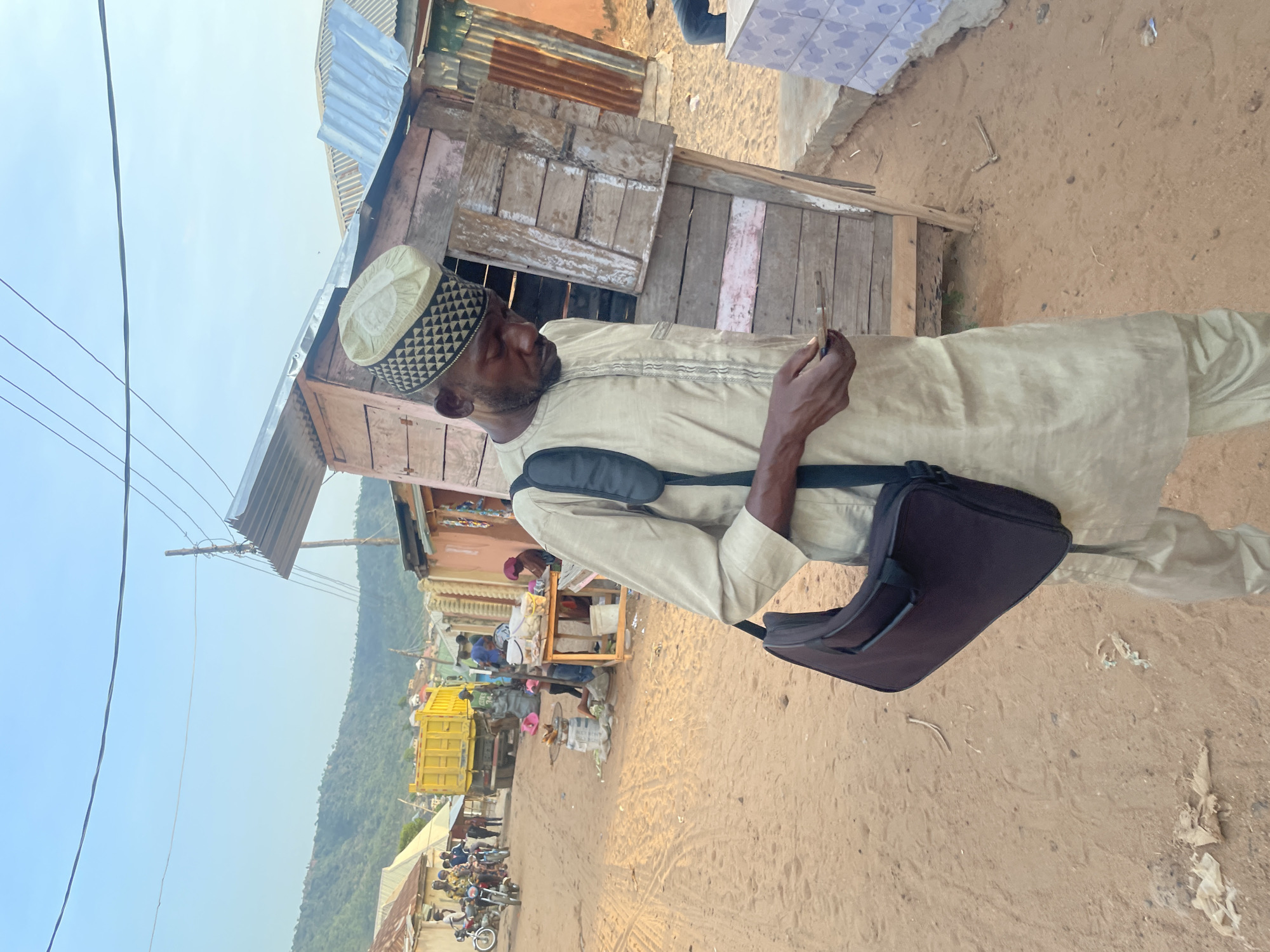 A man in a traditional cream-colored tunic and patterned cap stands outside a rustic wooden building in Lokoja, Nigeria. He has a black bag slung over his shoulder and appears to be examining something in his hand. In the background, a street scene unfolds with a yellow dump truck, people, motorcycles, and roadside vendors. A line of trees covers the horizon. The ground is sandy and uneven.