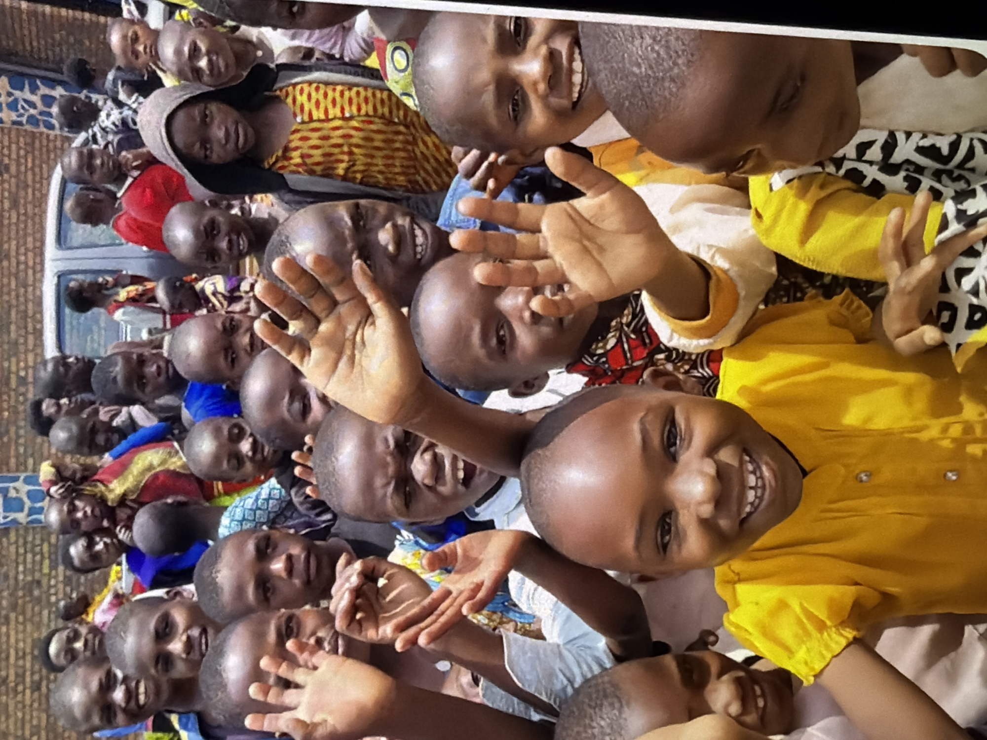 A group of children are gathered closely together, with many of them smiling and waving towards the camera. The children appear to be of African descent and are dressed in colorful clothing. In the background, a brick wall and a portion of a silver vehicle can be seen, suggesting an outdoor setting. The overall impression is one of joy and excitement.