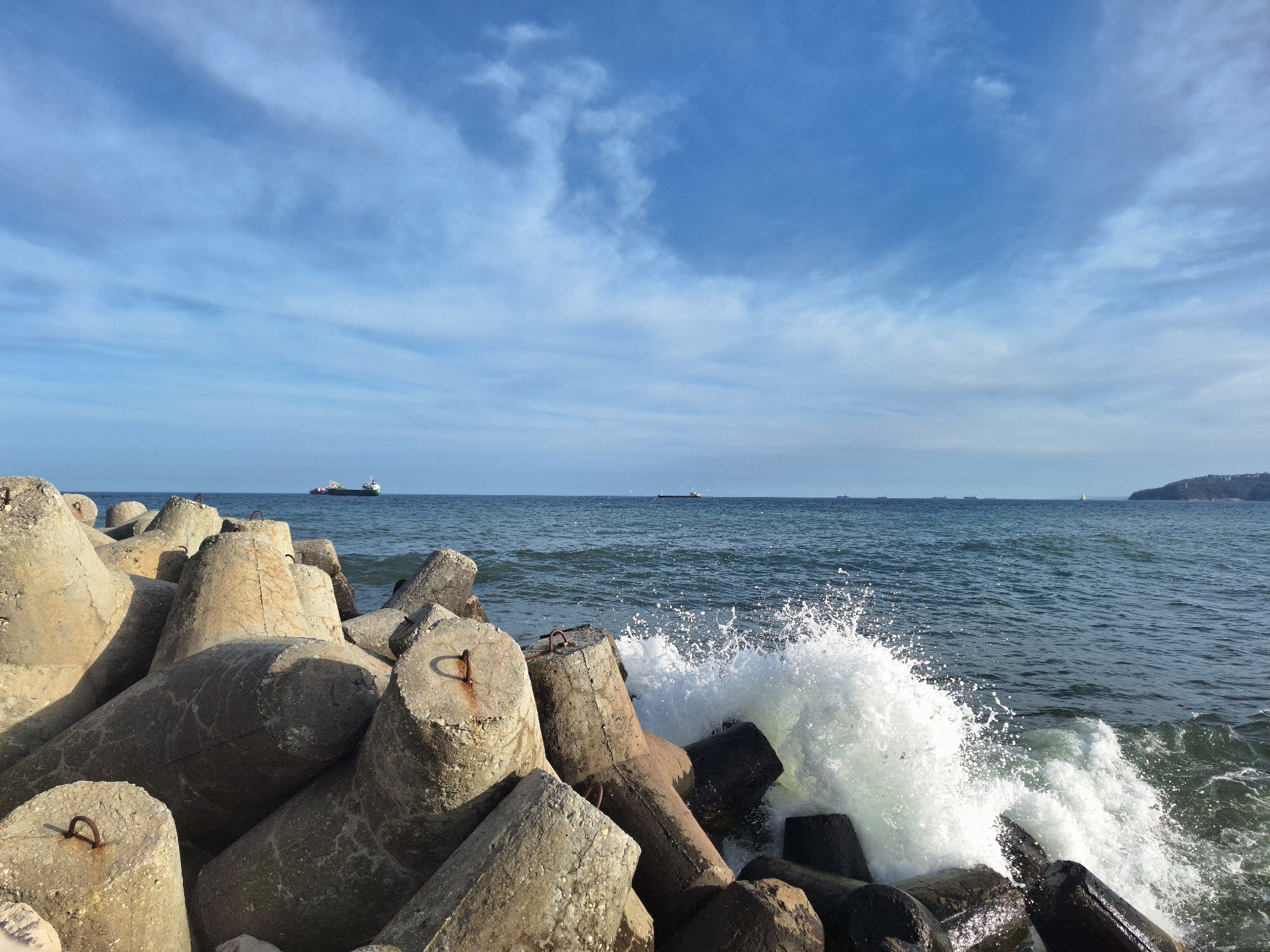 The image depicts a coastal scene in Varna, Bulgaria, with large concrete tetrapods forming a breakwater. Waves crash against the tetrapods, creating white splashes. In the background, the sea stretches to the horizon under a blue sky with scattered clouds. Several ships are visible in the distance. A headland with some buildings on top appears on the right horizon. The focus is on the interplay between the man-made structures and the natural forces of the sea.