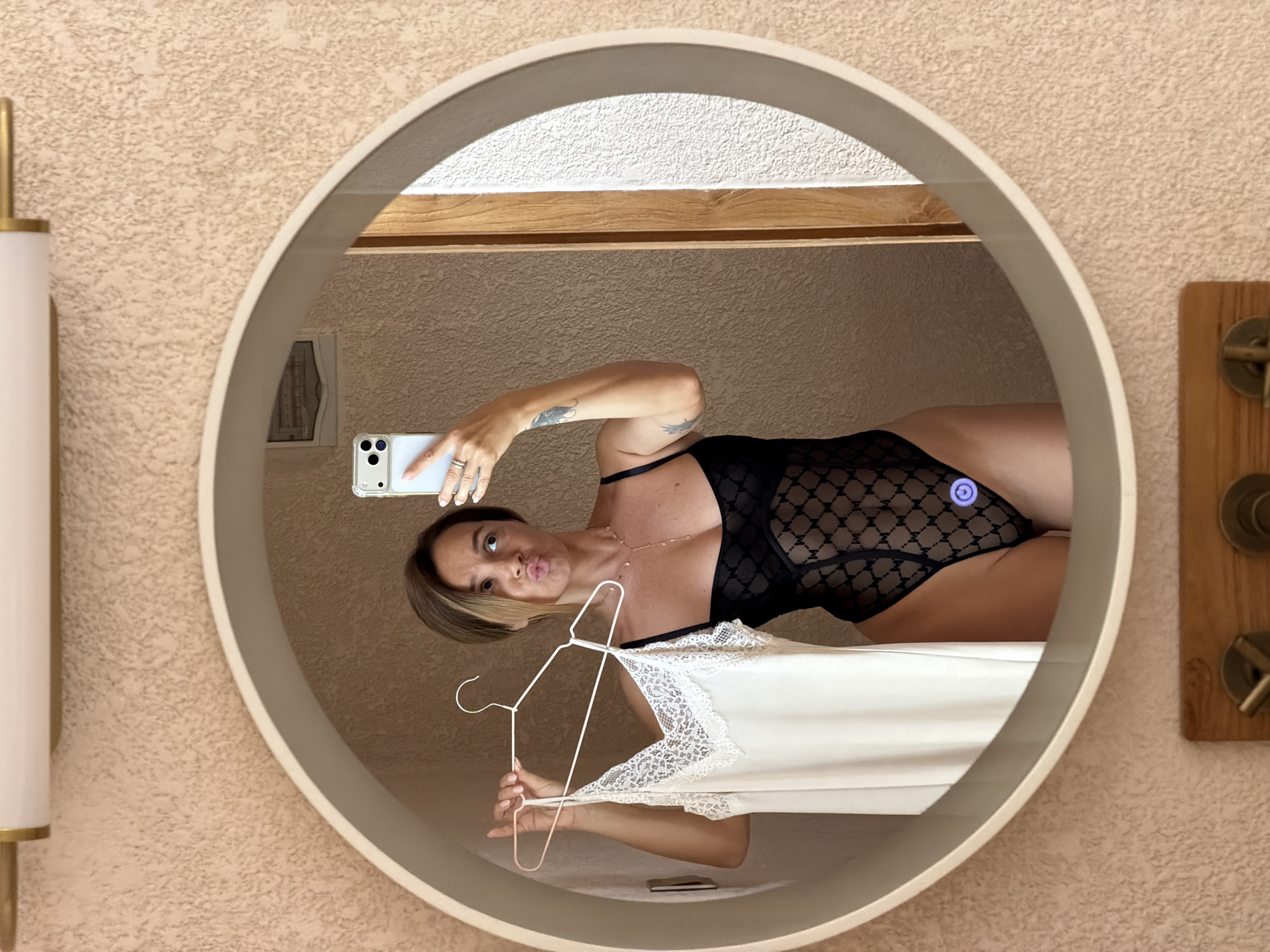 A woman is taking a mirror selfie in what appears to be a bathroom. She is wearing a black lace bodysuit and holding a white slip with lace trim on a hanger. The mirror is round and backlit, and she is making a kissing face. The background is a textured pink wall. The photo is taken from the chest up, and the woman has short, brown hair. There are two light fixtures visible above the mirror, and what appears to be a wooden door frame on the right side of the image. There is also a small blue circular light on the bodysuit.